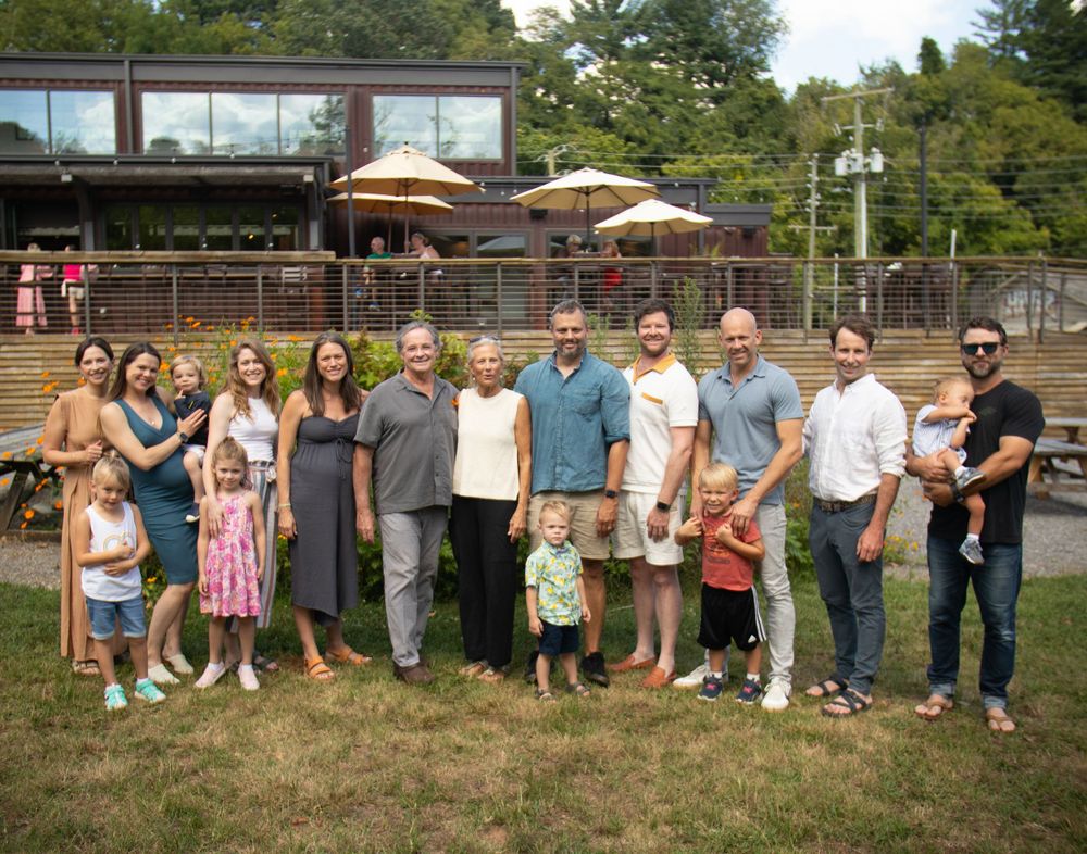 Large group gathers outdoors for casual family photo in front of restaurant patio on sunny day.