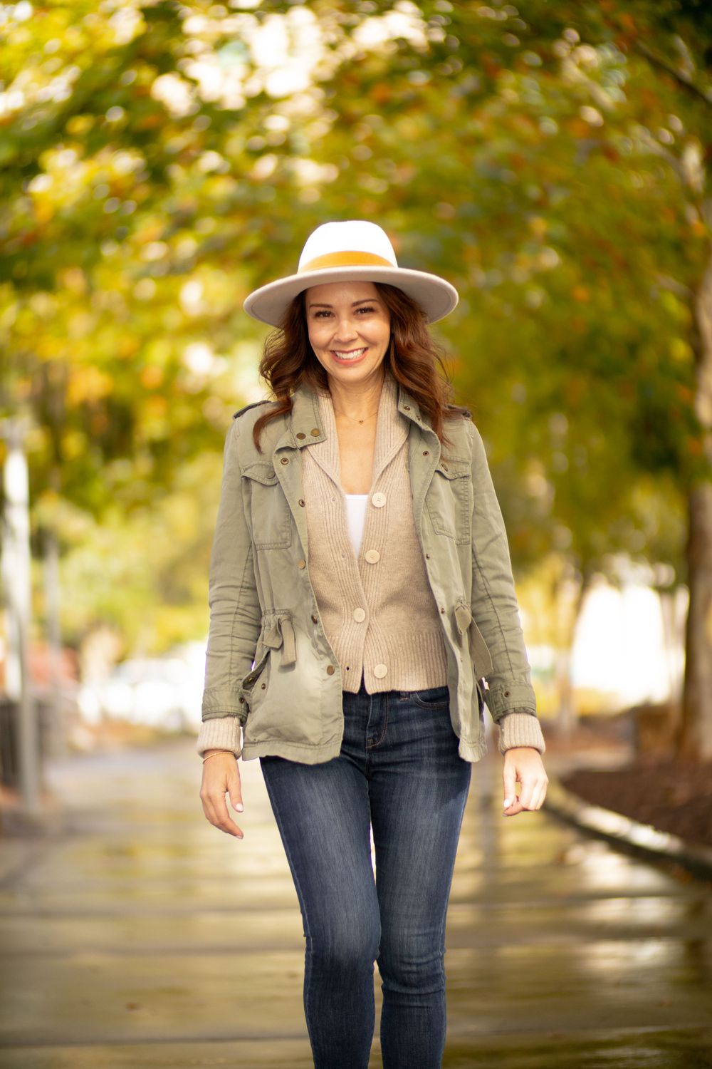 Fall fashion look with neutral fedora, olive utility jacket and fitted jeans against autumn foliage.