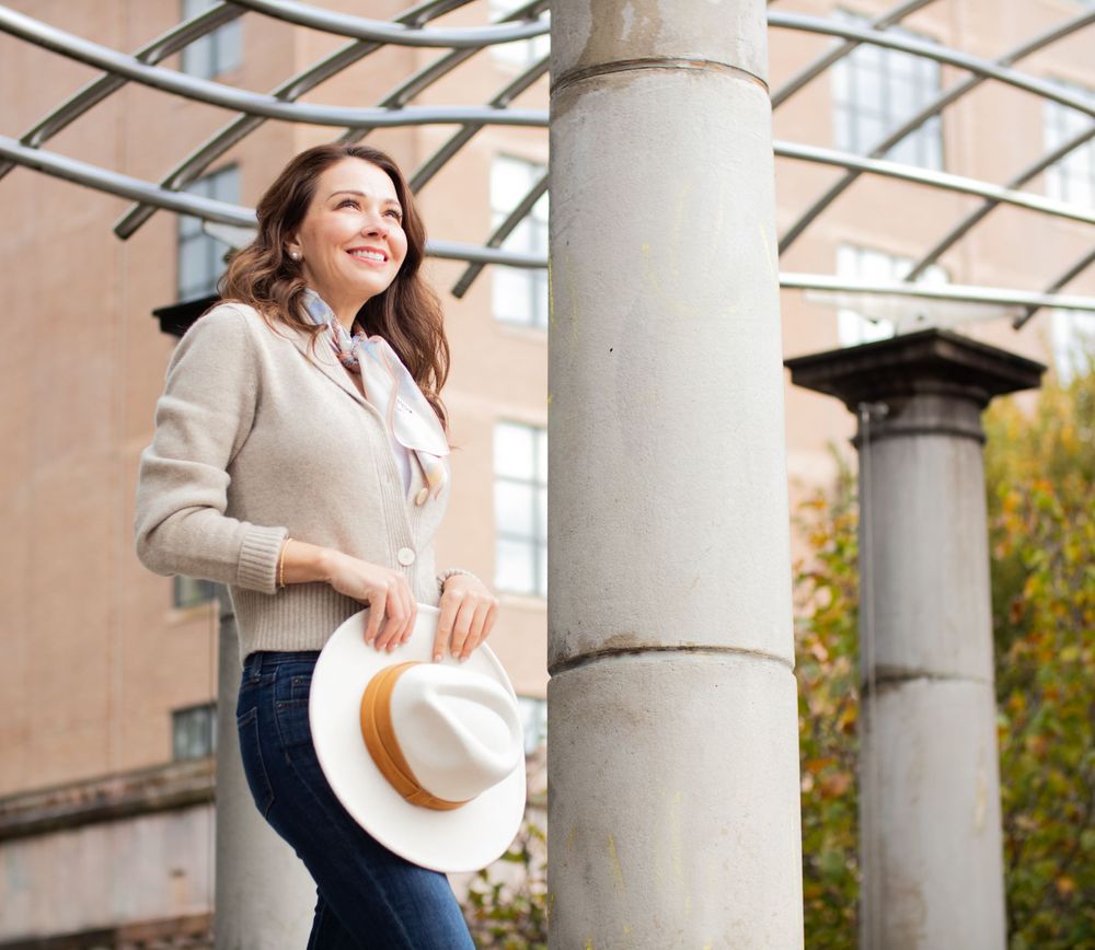Candid lifestyle photo near columns wearing beige sweater and holding cream fedora.