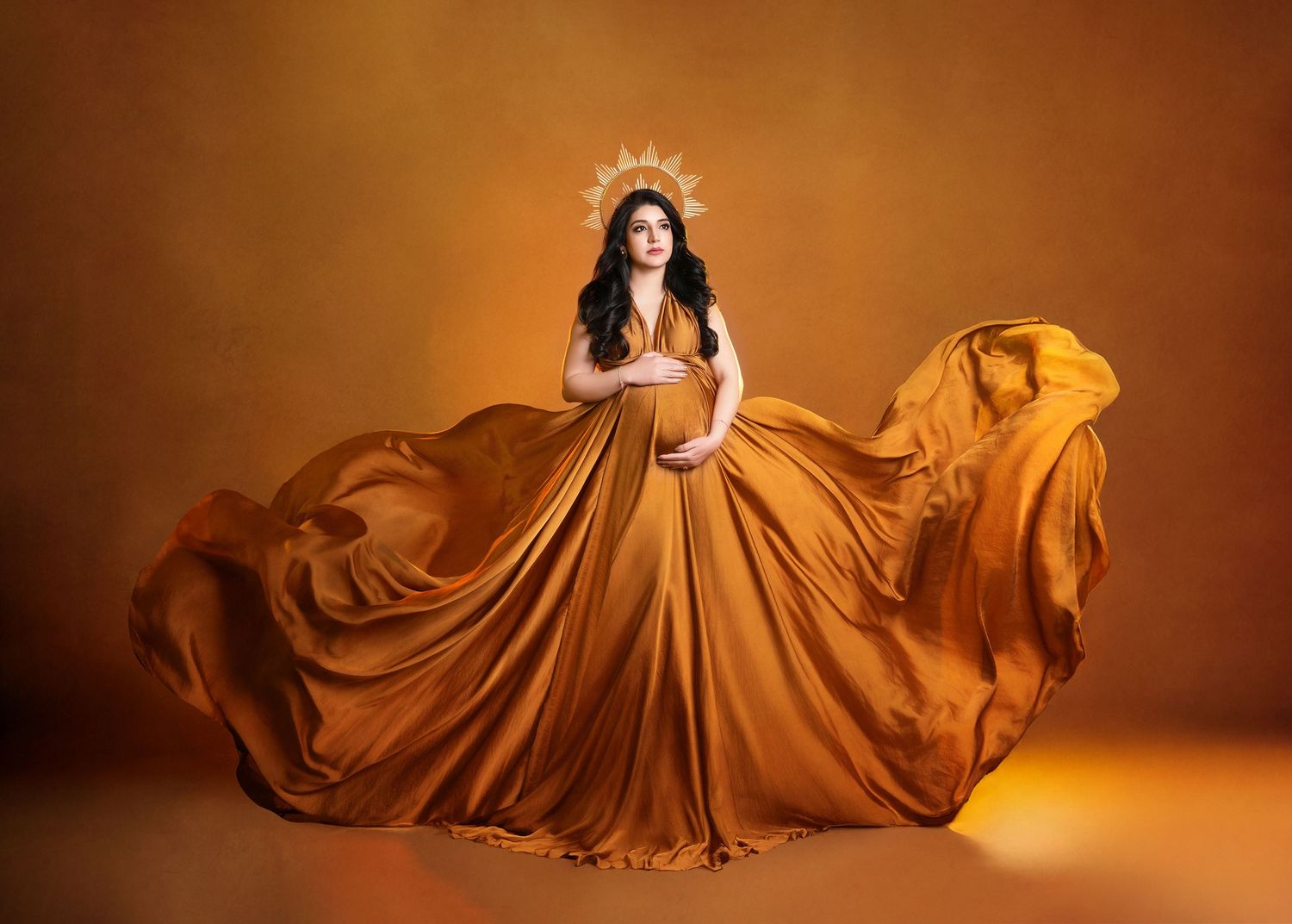 A figure in a flowing caramel-colored gown and crown poses dramatically against a warm brown studio backdrop.
