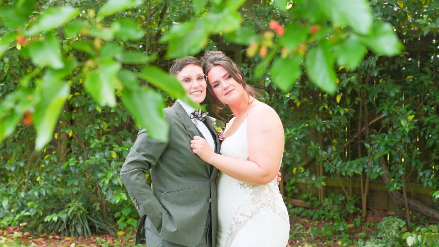 A couple poses together for a wedding photo outdoors among green foliage on their special day.