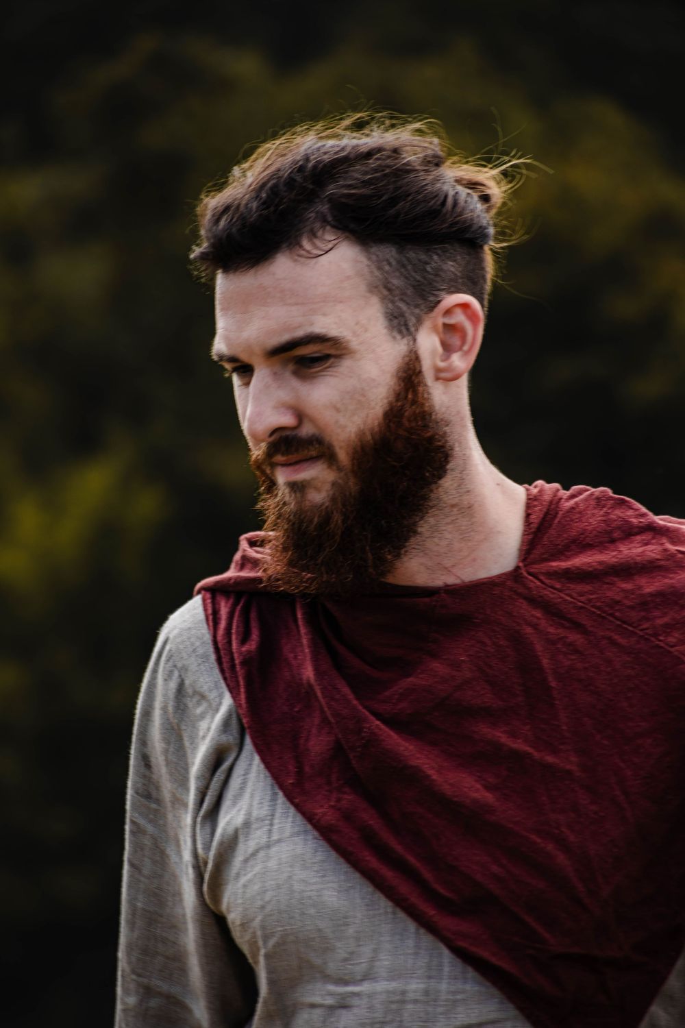Portrait of a person wearing a burgundy cloak and gray shirt with windswept hair against a dark background.
