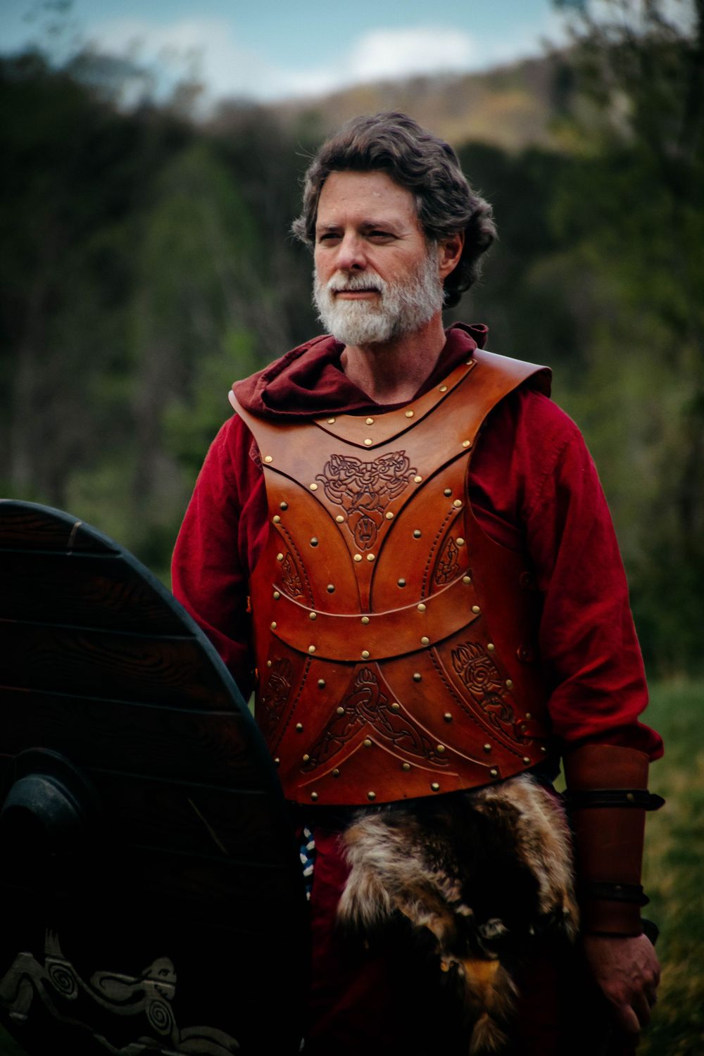LARP enthusiast wearing a red medieval tunic and detailed brown leather armor poses against a nature backdrop.