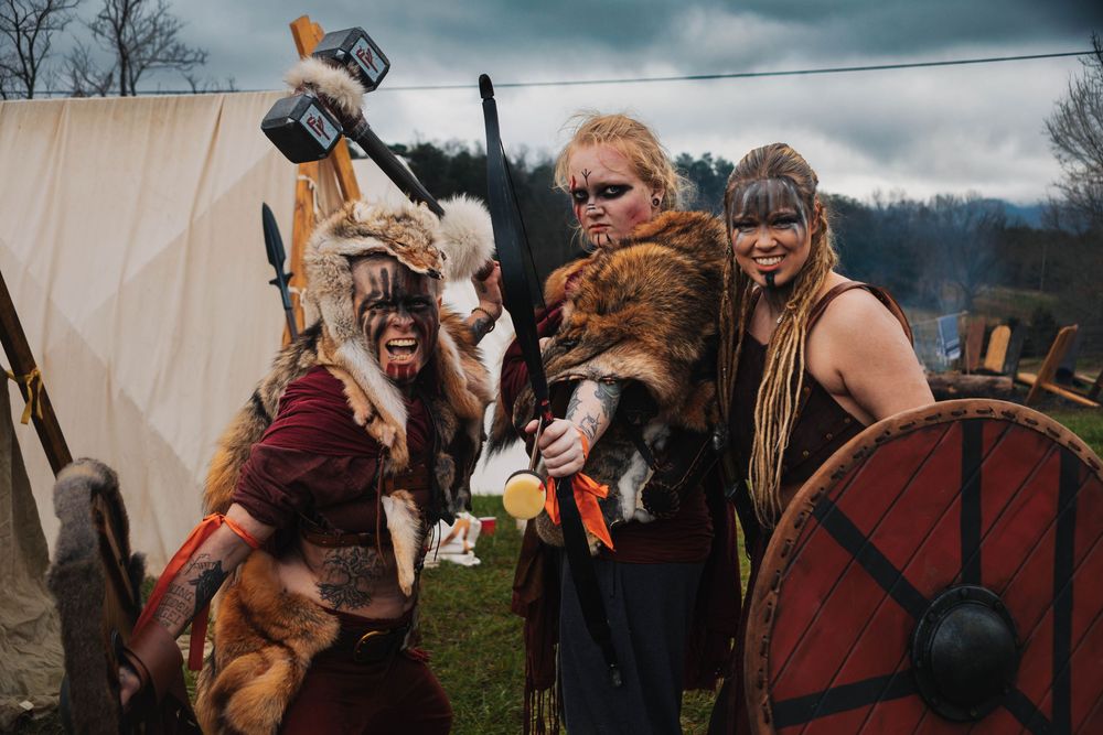 People in Viking costumes pose with fur pelts and weapons at an outdoor reenactment event against cloudy sky.