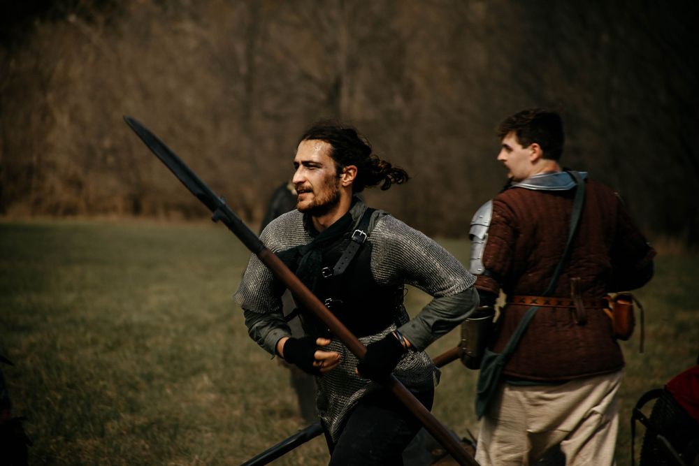 Two medieval warriors in period costumes practice with weapons in a grassy field during combat training.