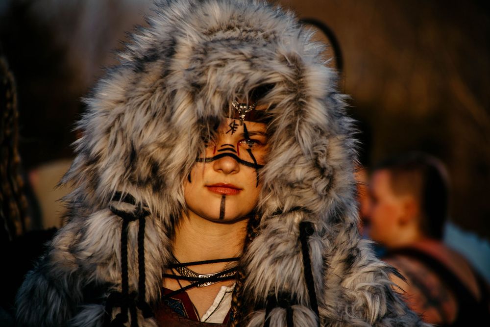 Person wearing winter parka with fur-lined hood and face paint illuminated by warm orange light at dusk.