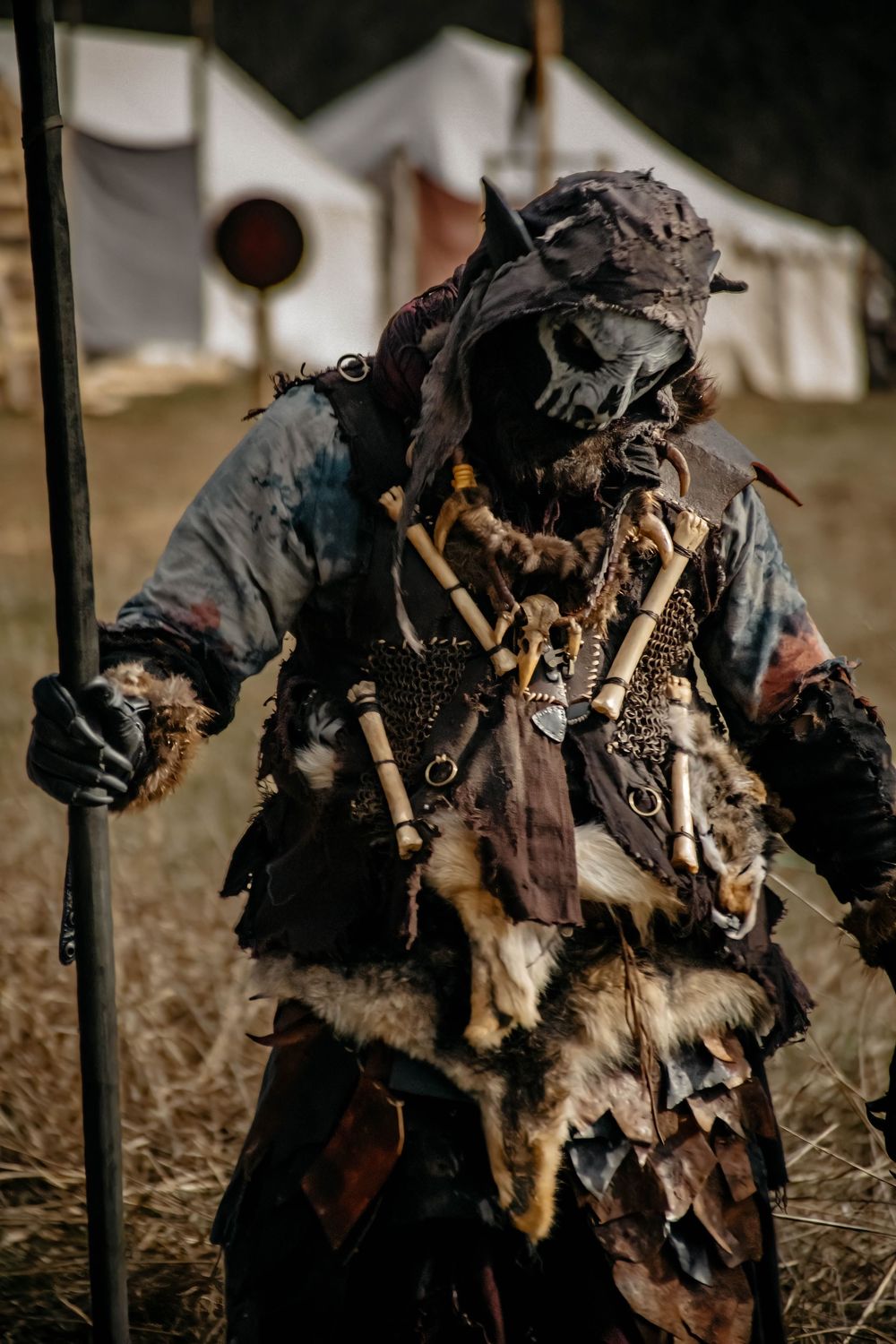 Costumed figure in post-apocalyptic wasteland attire with weathered clothing and decorative mask holds staff in outdoor setting.