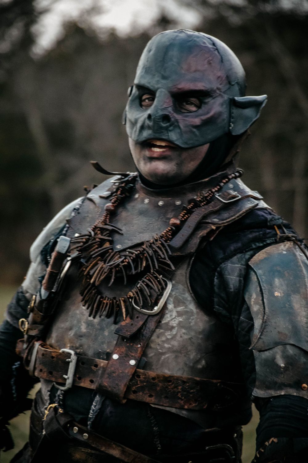 A cosplay character wearing detailed fantasy orc armor with leather straps and blue-gray prosthetic makeup in a dark setting.