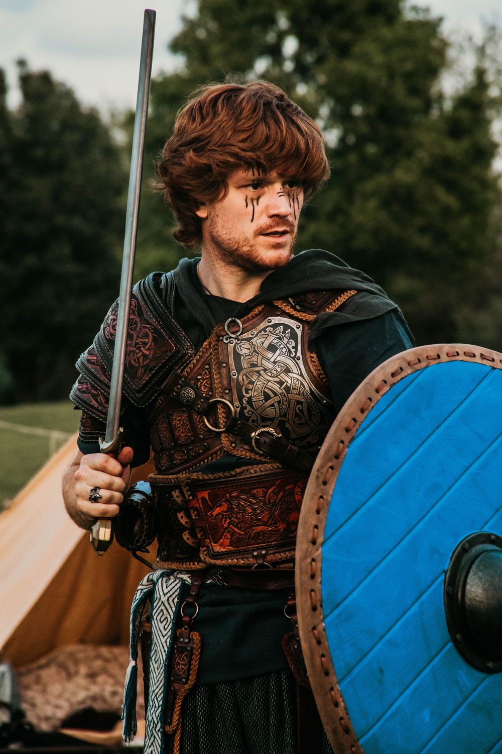 A medieval warrior in dark armor and leather stands ready with a sword and bright blue wooden shield.