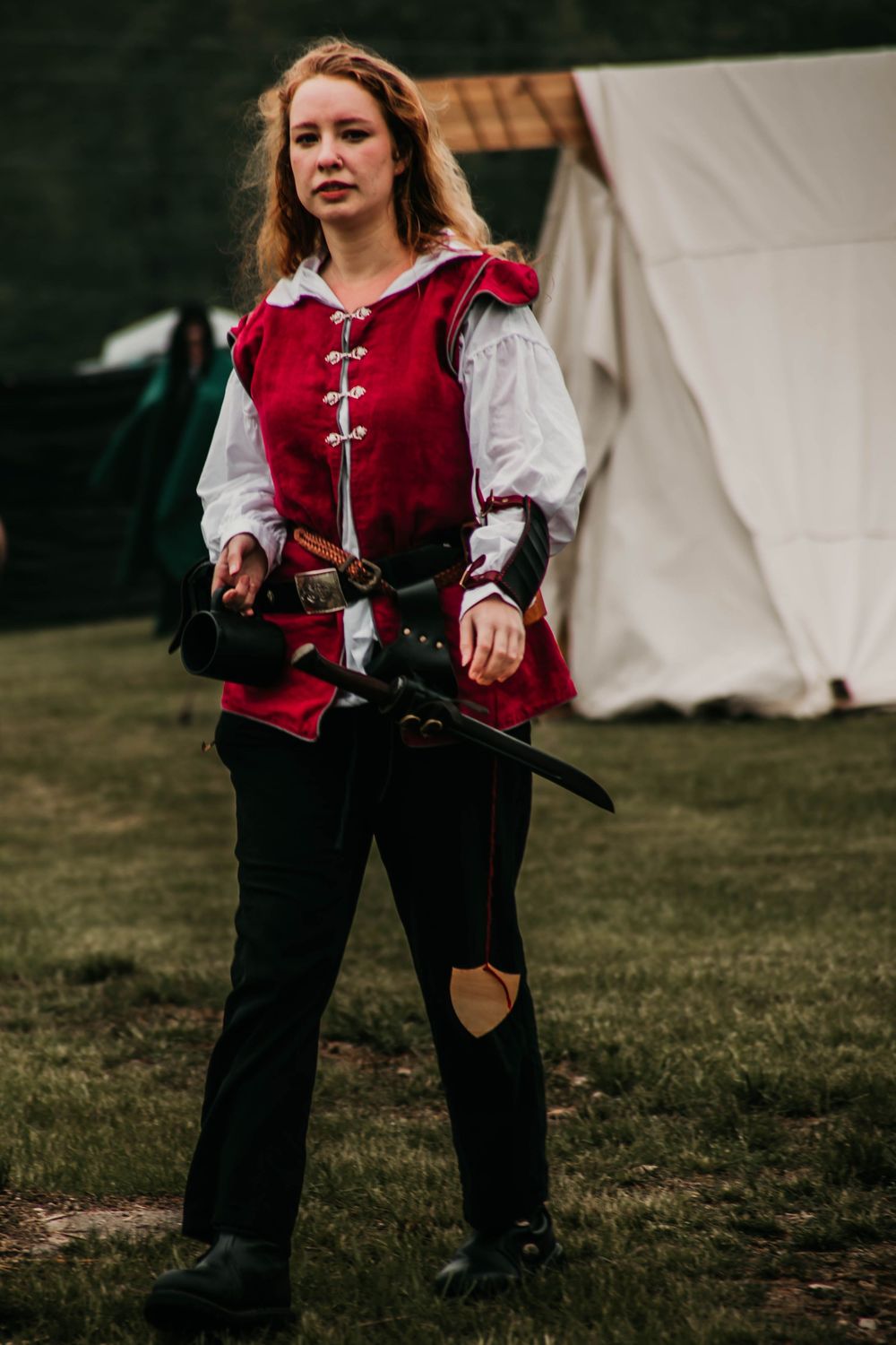 A person wearing a medieval-style red and white costume with black pants stands near canvas tents at a historical reenactment.