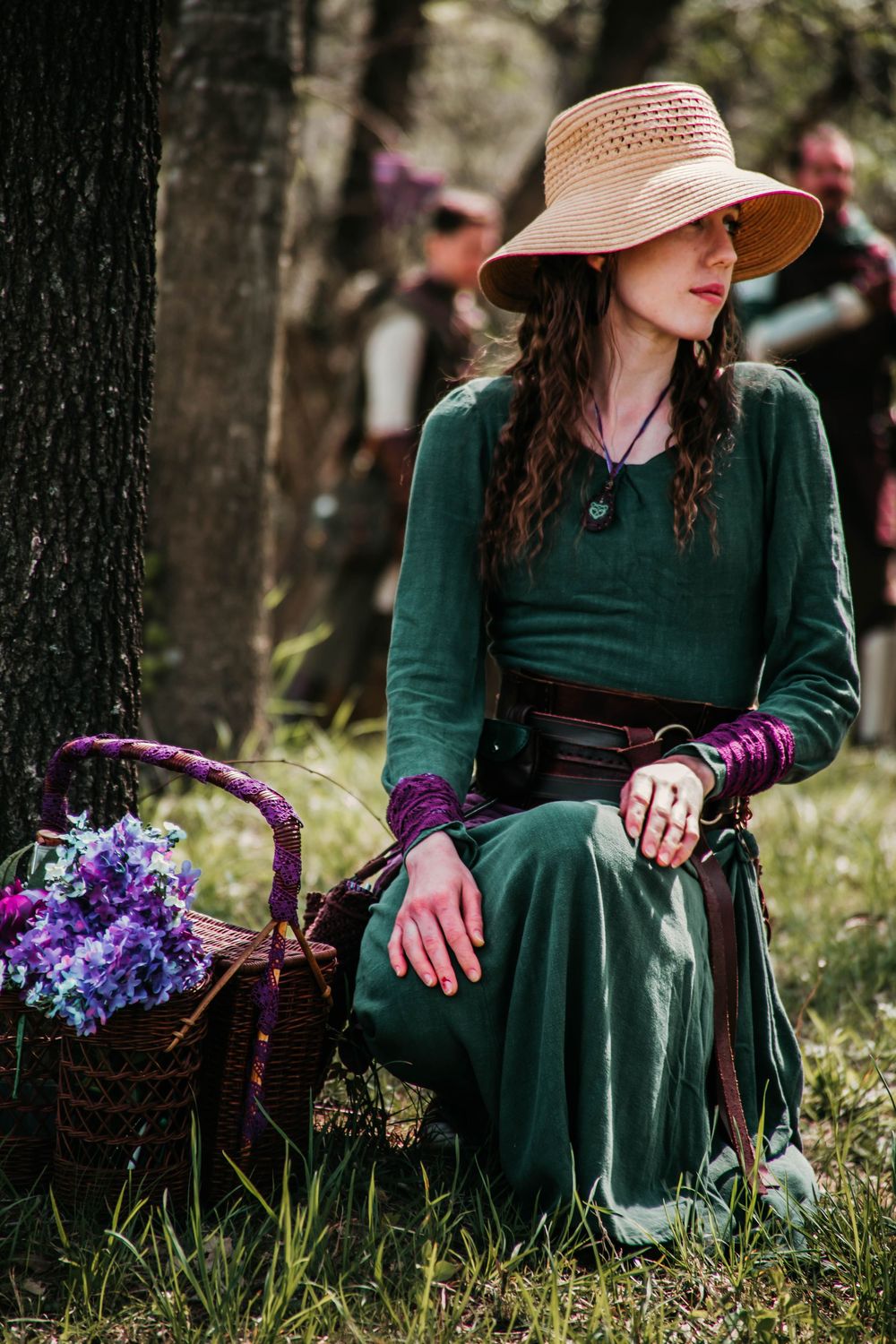 Model wearing green dress and straw hat sits by tree with purple flowers in wicker basket.