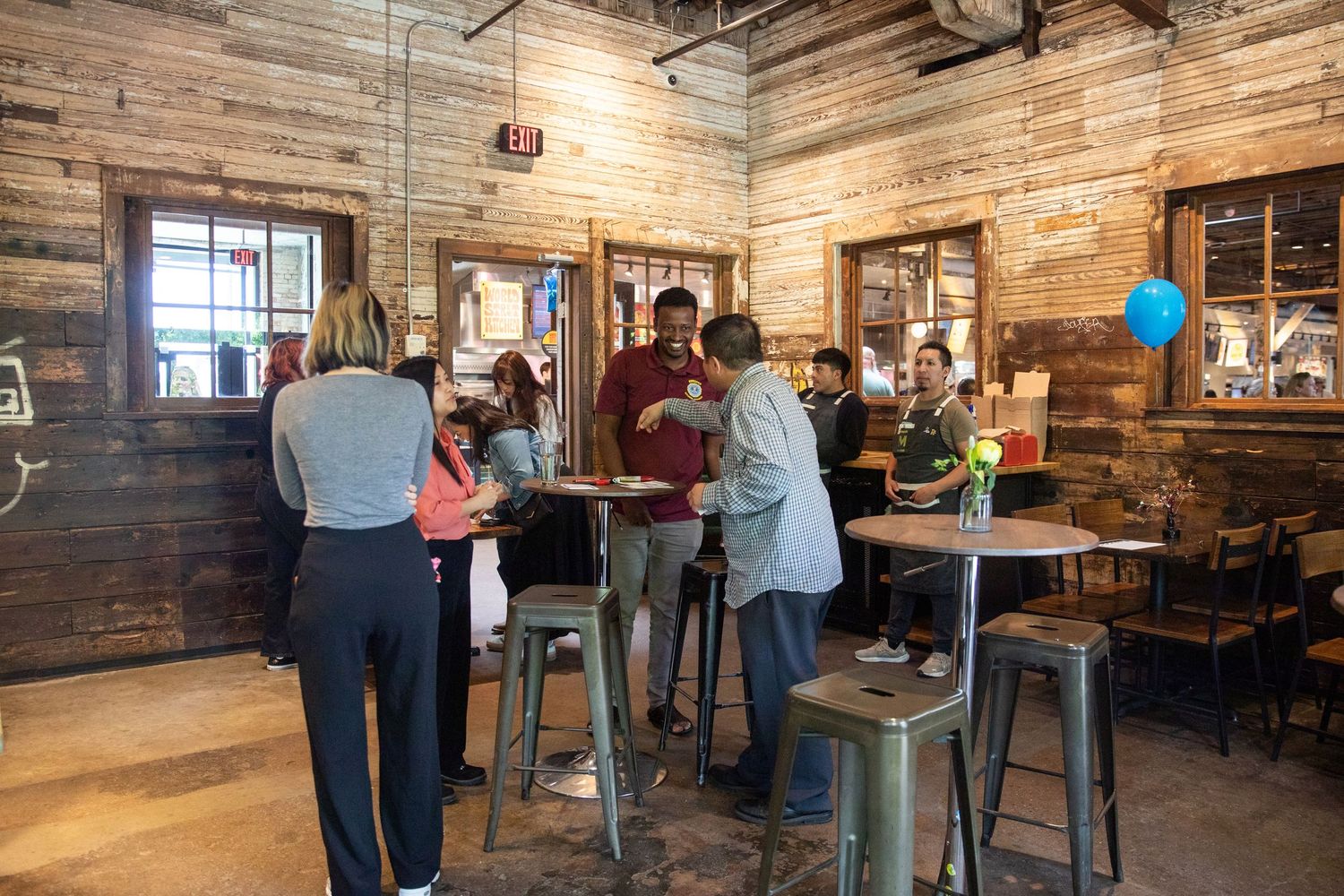 People gather at high-top tables inside a rustic bar with wooden walls and industrial lighting.