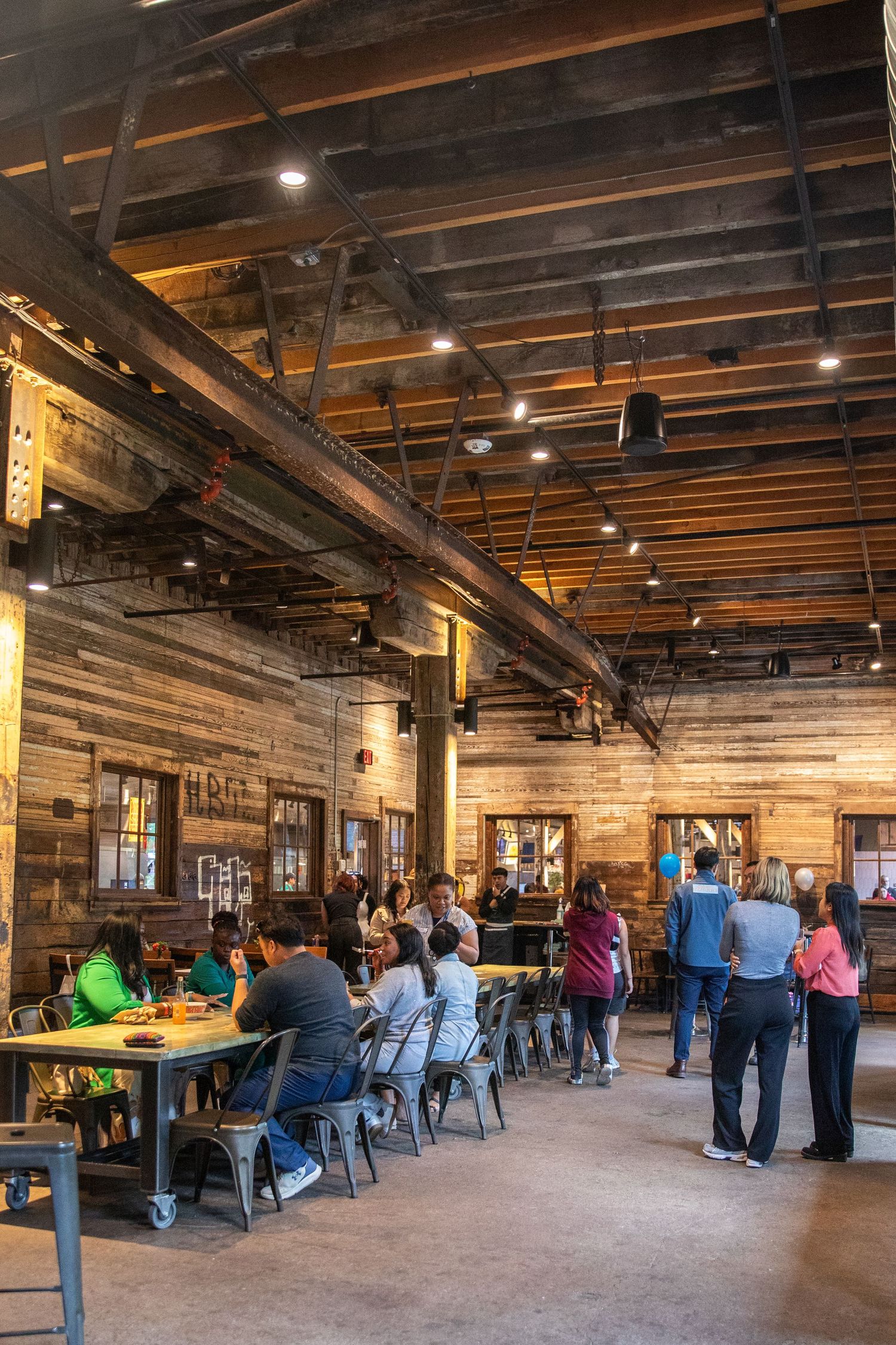 Rustic restaurant interior with wooden walls, industrial ceiling beams, and diners seated at communal tables.