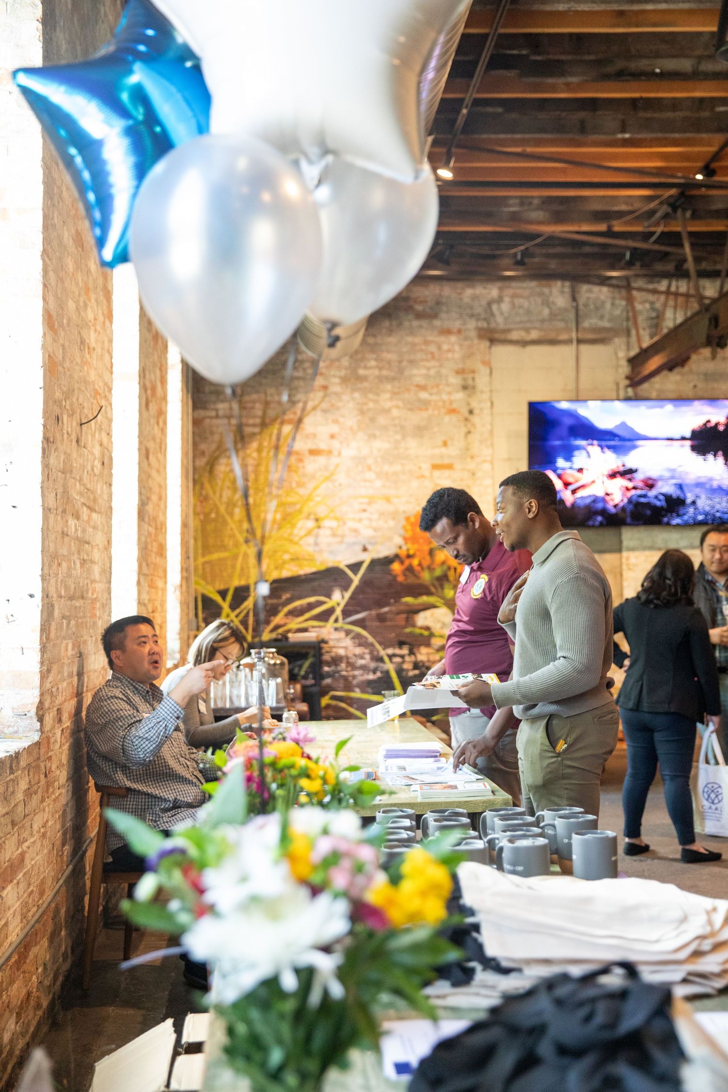 People gather around a buffet setup with catering trays at an event decorated with blue and white balloons.