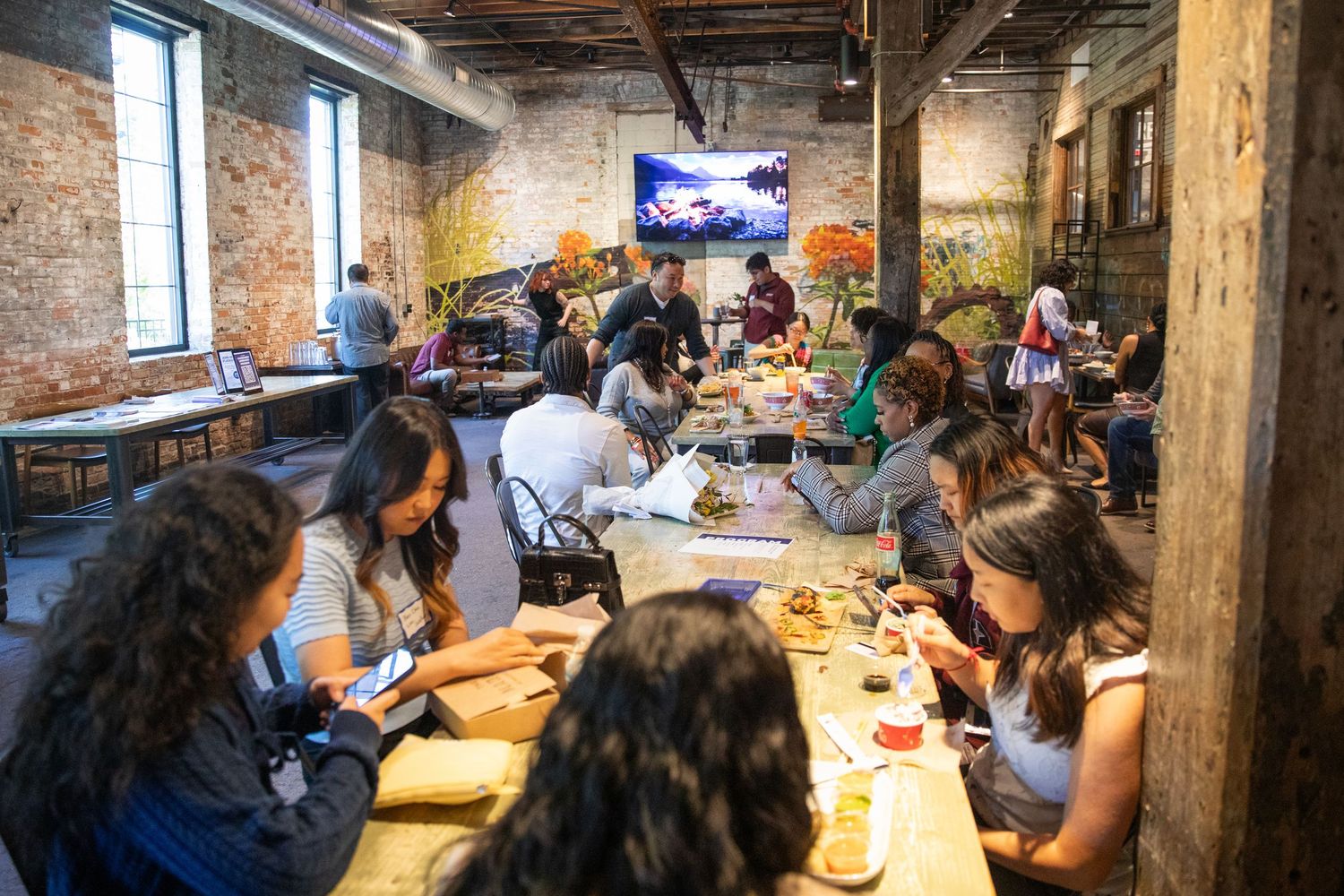 A group gathers for a meal at wooden tables inside a rustic brick-walled restaurant with exposed beams and windows.