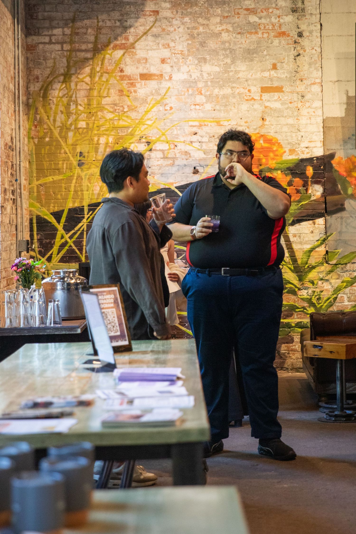 Two restaurant workers having a conversation in front of an exposed brick wall with greenery and table setup.