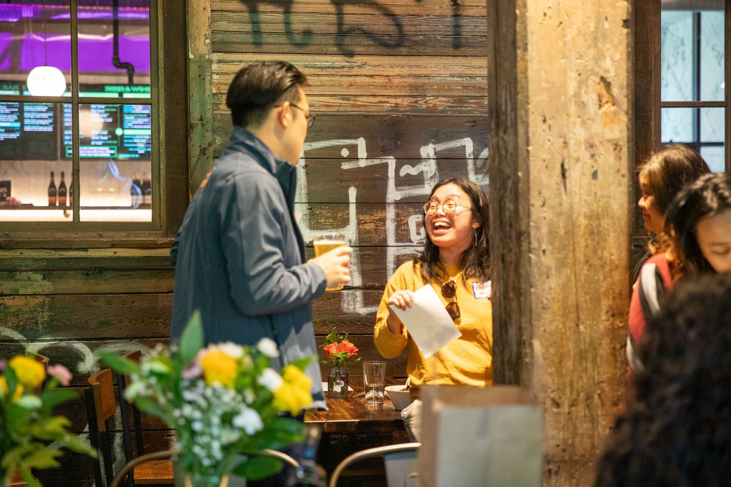 Two people having an animated conversation at a rustic cafe with brick walls and colorful flowers in the foreground.