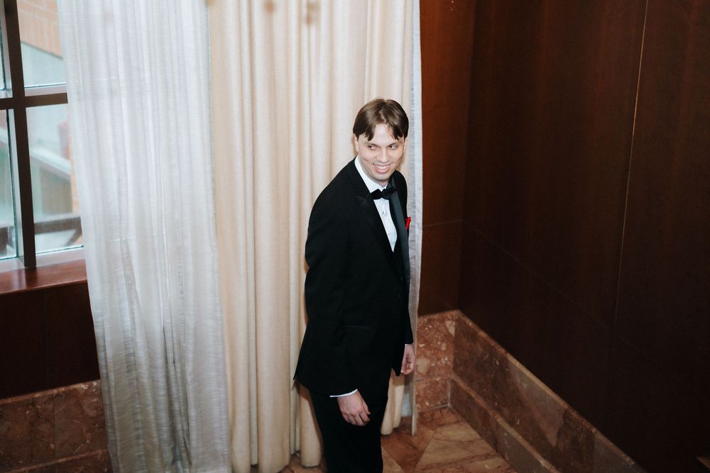 Men in formal attire walk through an elegant wood-paneled hallway with chandeliers.