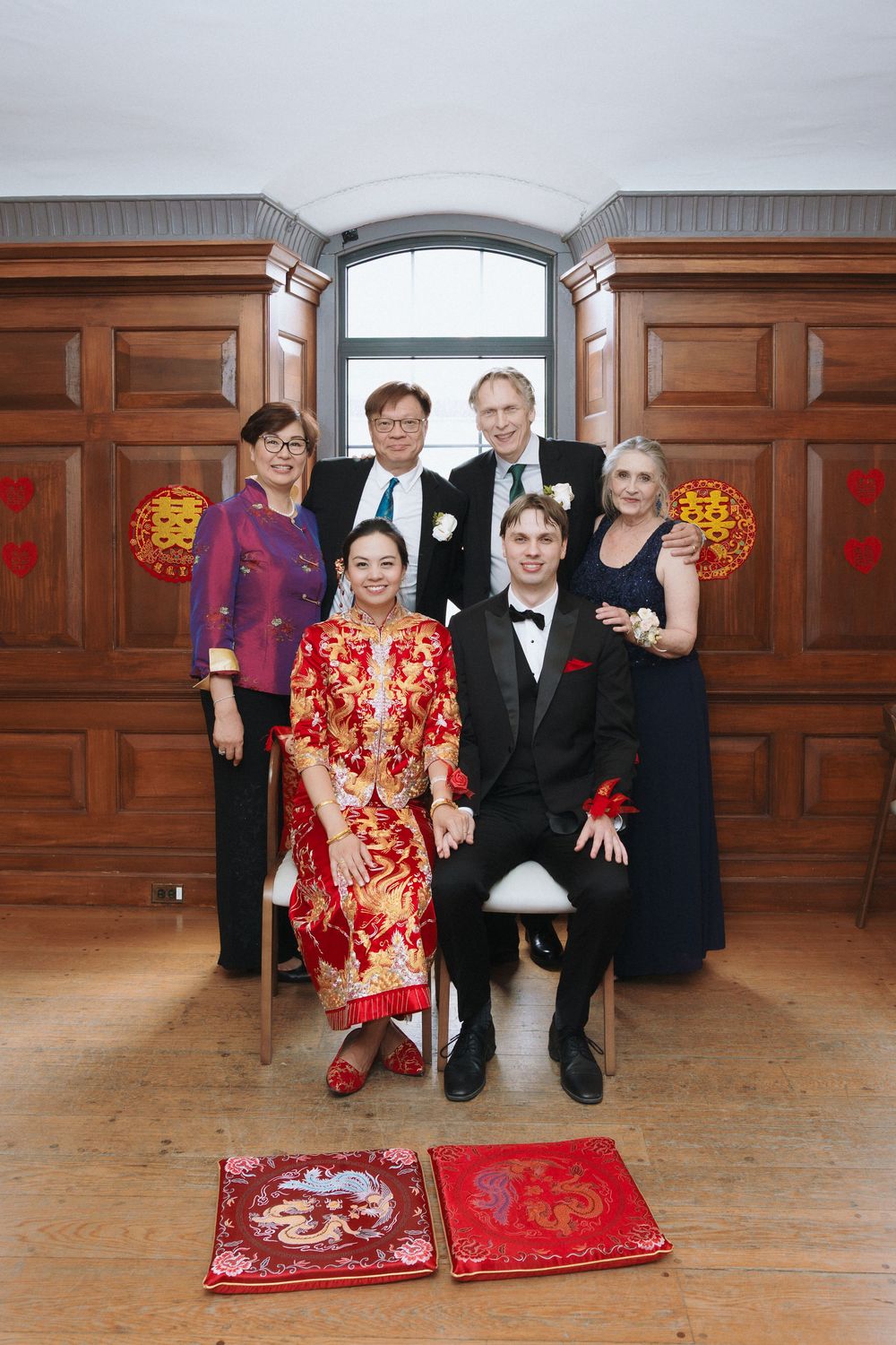 Family wedding portrait featuring traditional Chinese red dress and Western formal attire in elegant wood-paneled room.