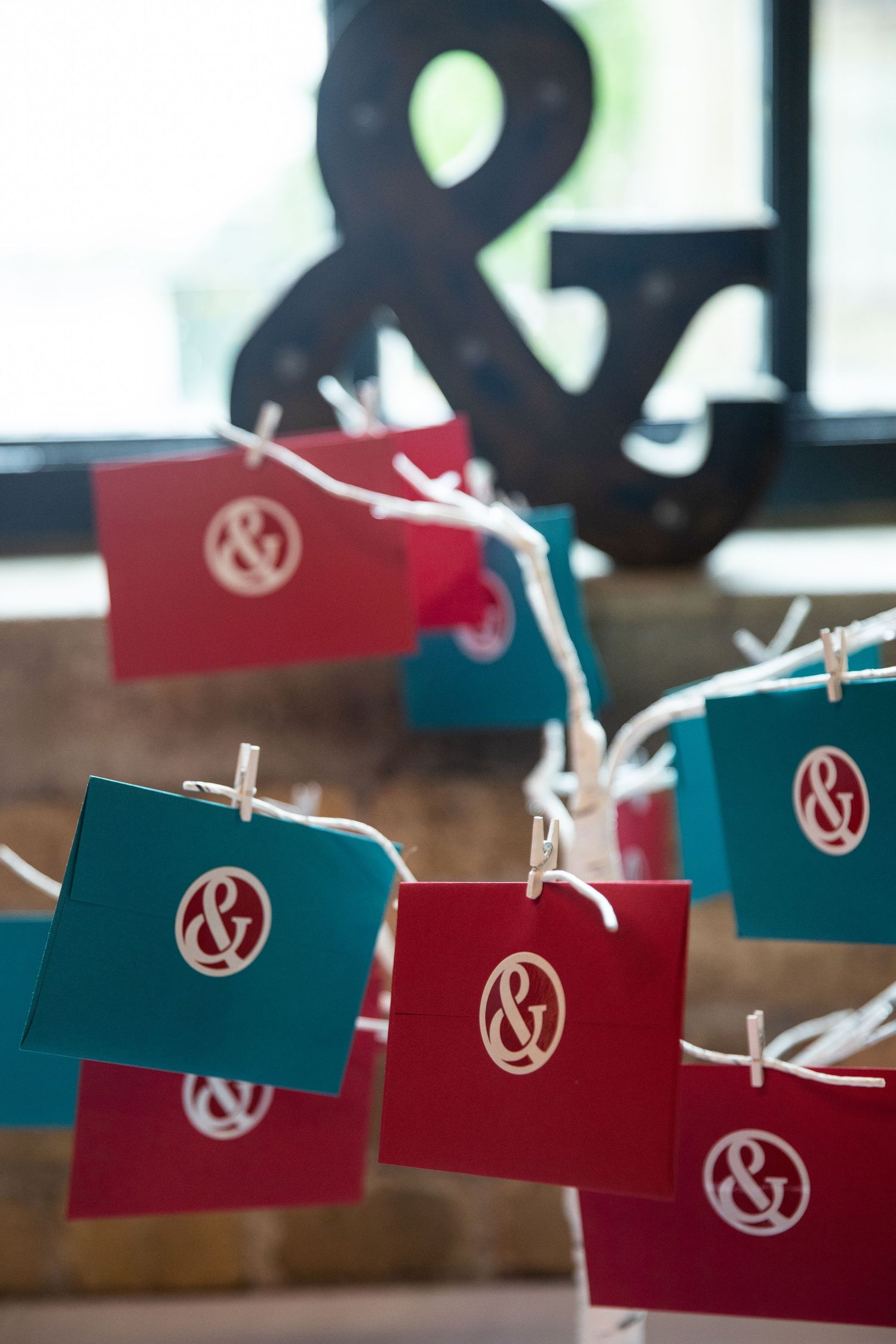 Small red and teal paper cards with ampersand logos hanging on a string with clothespins against a blurred background.