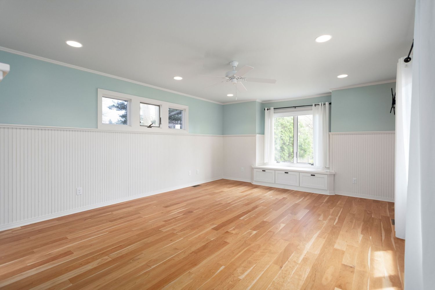 Empty room with light blue walls, white beadboard wainscoting, hardwood floors and natural light from a window.