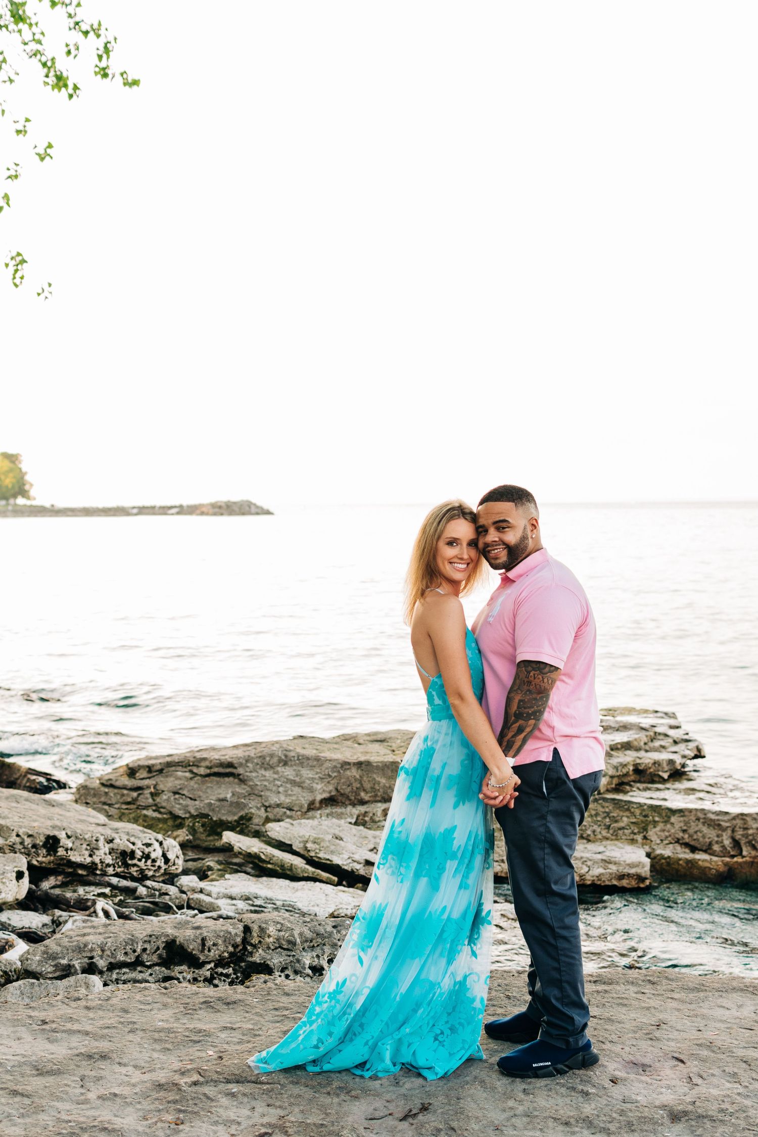 A couple embraces on rocky coastal shore at sunset, with the woman wearing a flowing turquoise dress.
