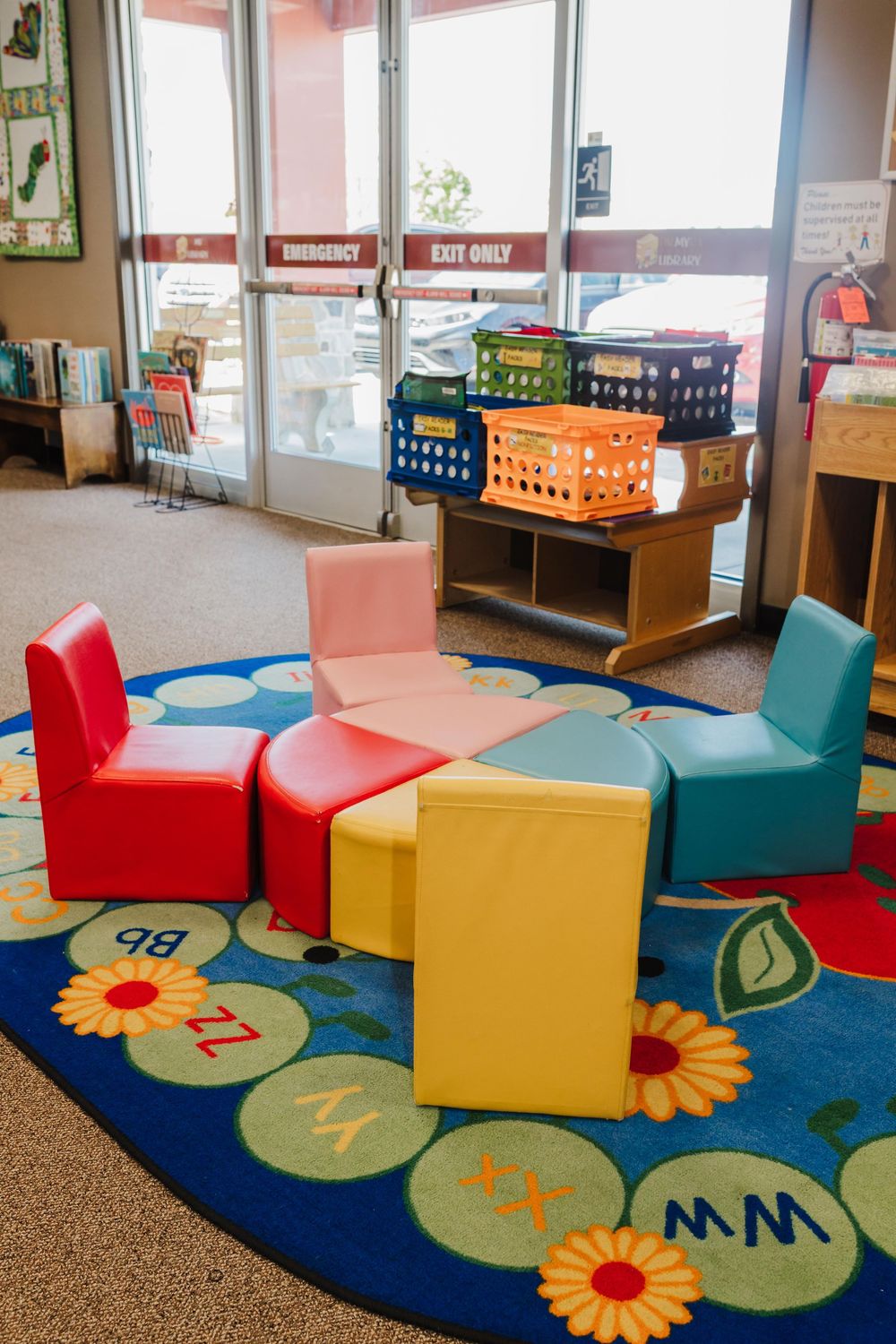 Colorful kid-sized chairs arranged in a circle on a floral educational rug with storage baskets by the windows.