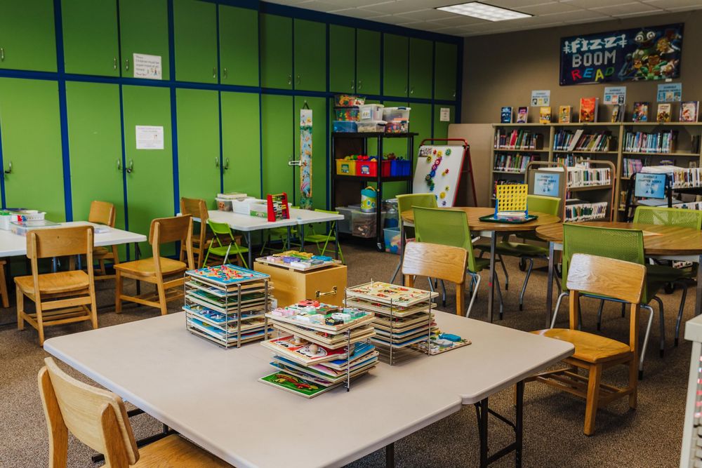 A library room with bright green walls, wooden chairs, and stacks of books, puzzles, and games on white tables near bookshelves.