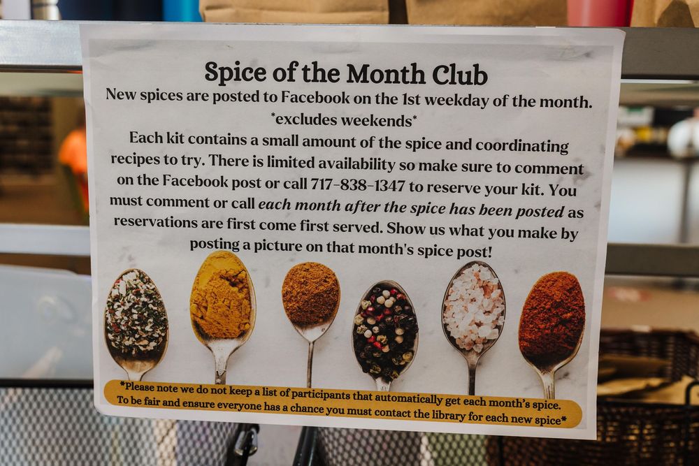 A sign displays five different colored spices for a "Spice of the Month" club with details on how to participate.
