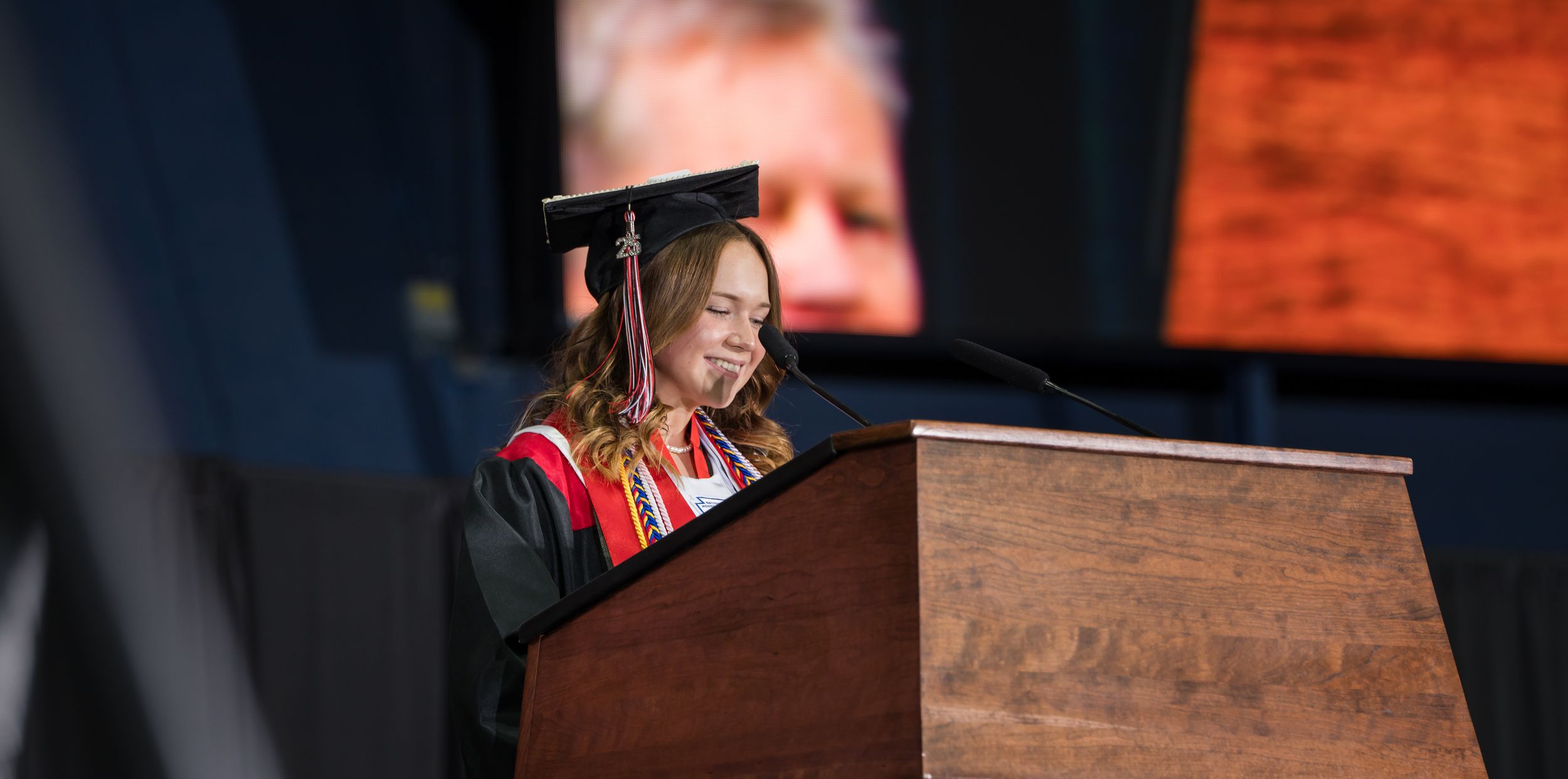 Graduate stands at podium decorated with colorful flowers during outdoor commencement ceremony.