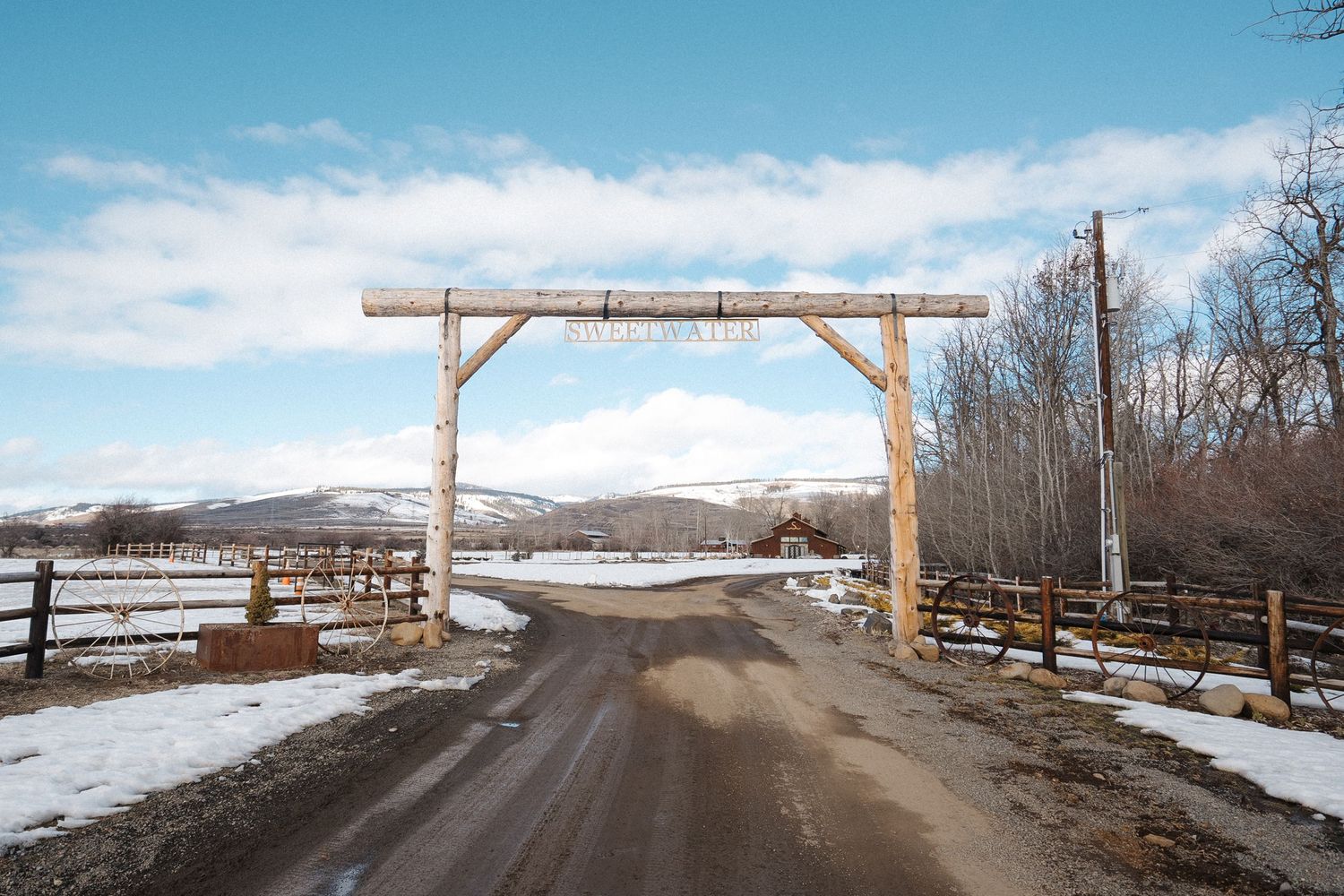 A wooden ranch entrance gate spans a snowy dirt road with wooden fences and mountains in the background.