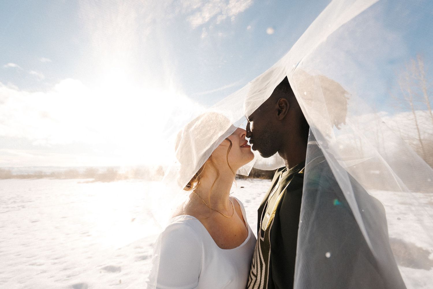 A couple in wedding attire under a flowing veil share an intimate moment in a bright outdoor setting with white landscape.
