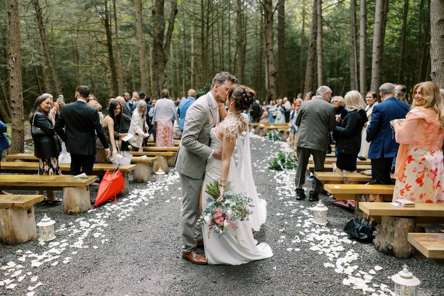 A couple shares their first kiss on a flower petal covered forest path during an outdoor ceremony with wooden benching at Windham Manor.
