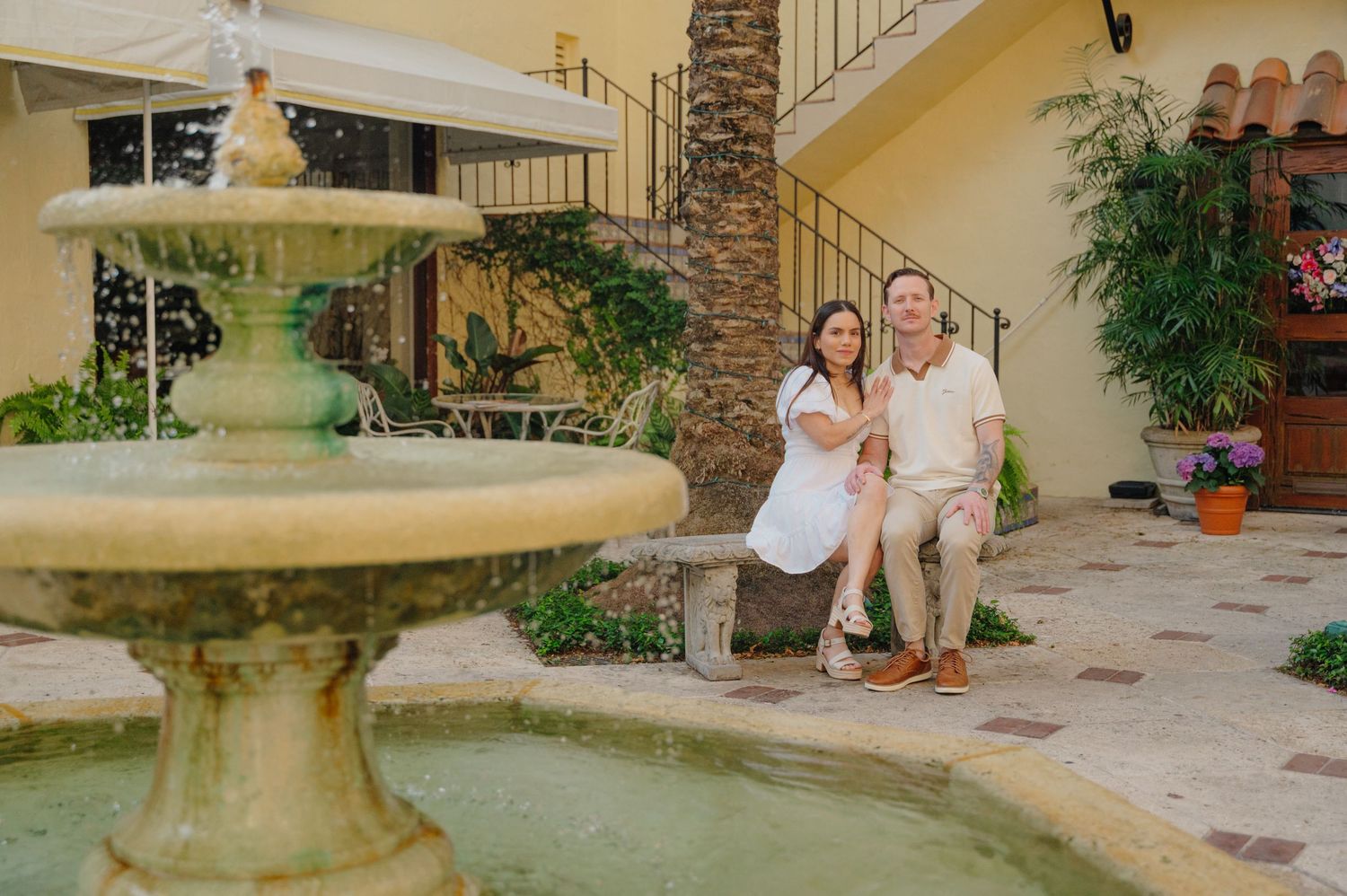 A stone fountain stands in front of a Mediterranean-style home with terracotta pots and climbing vines on the patio.