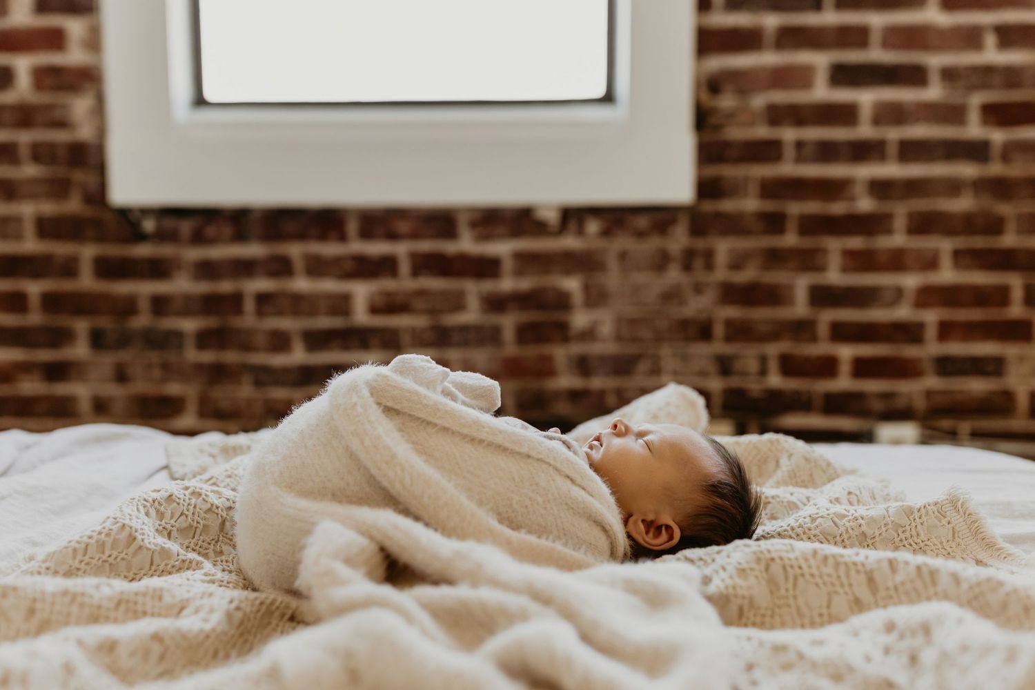 Someone relaxes peacefully on a cozy bed against an exposed brick wall background. Tulsa newborn photographer
