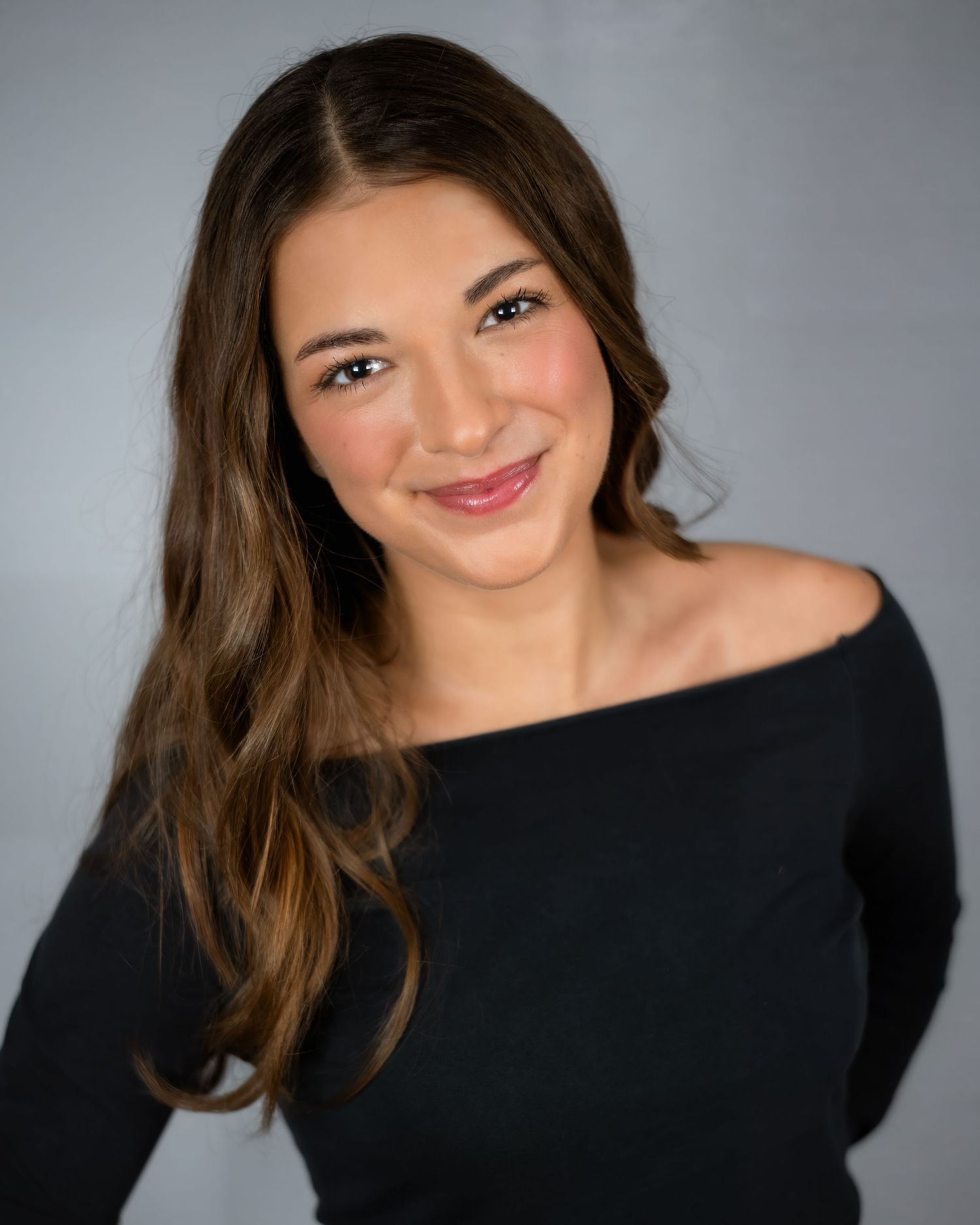 A professional headshot of a confident person wearing a stylish black off-shoulder top against a gray background.