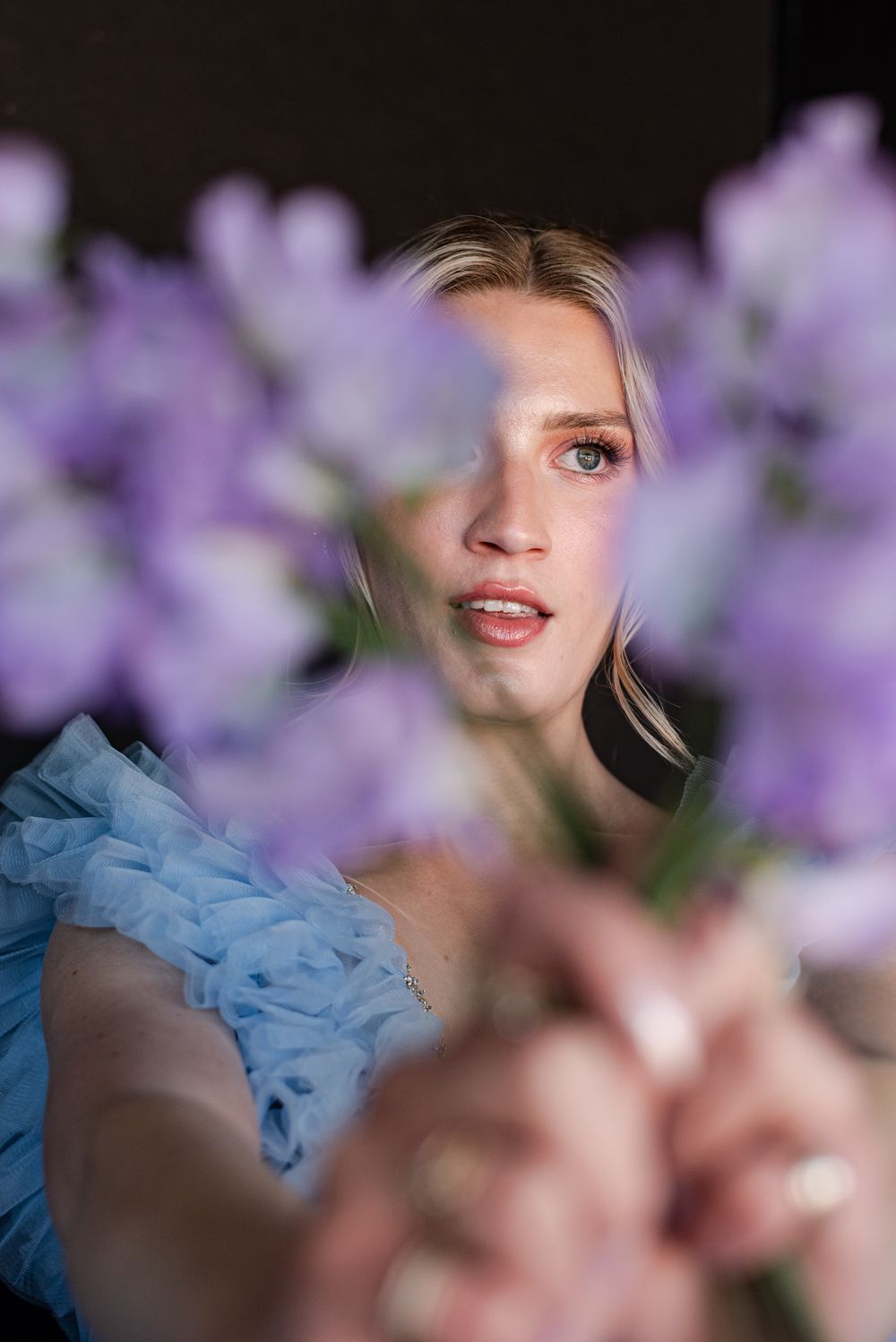 Ethereal portrait through purple flowers with soft focus showing a light blue tulle dress in dreamy lighting.