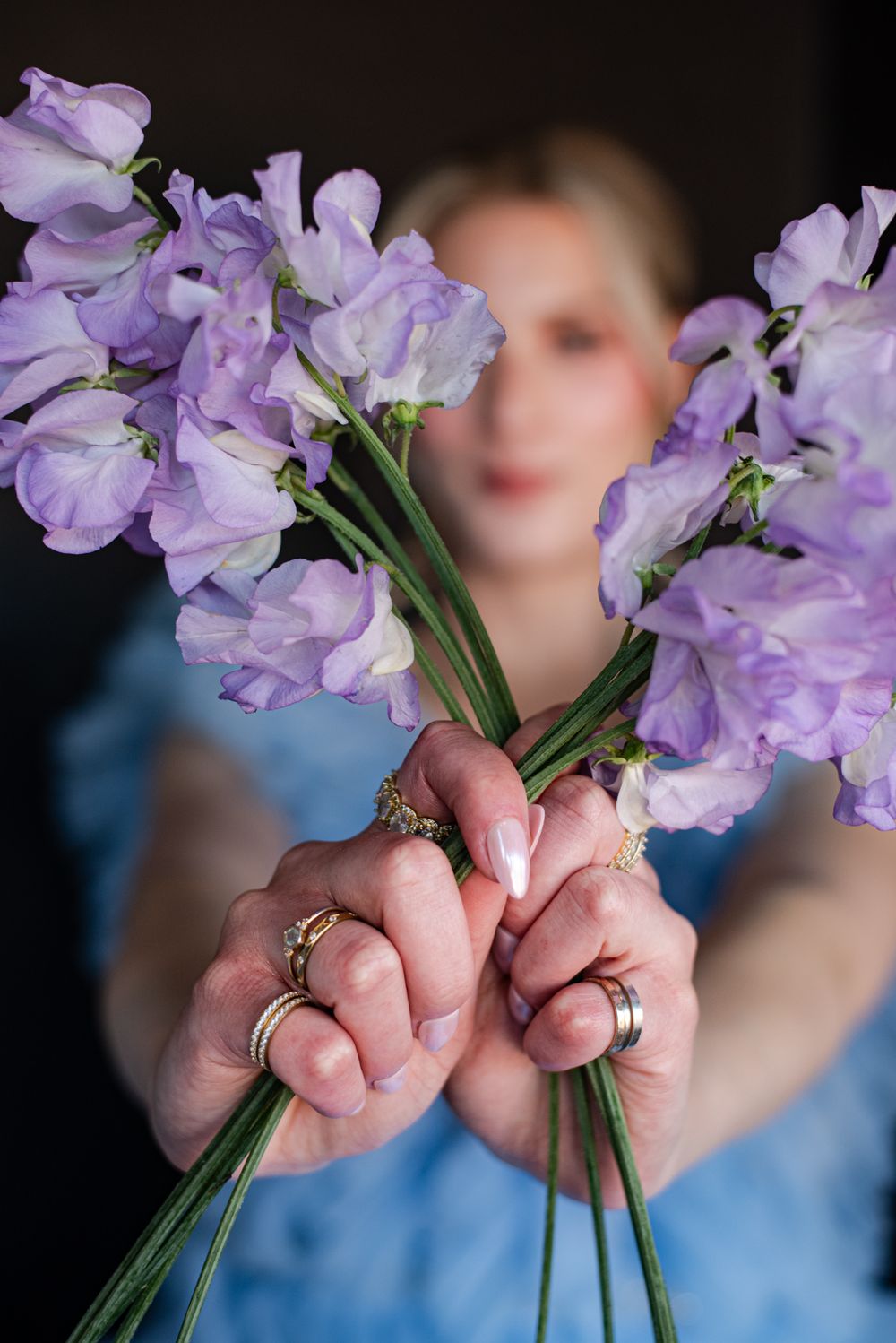 Hands adorned with rings hold delicate purple sweet pea flowers against a blurred background.