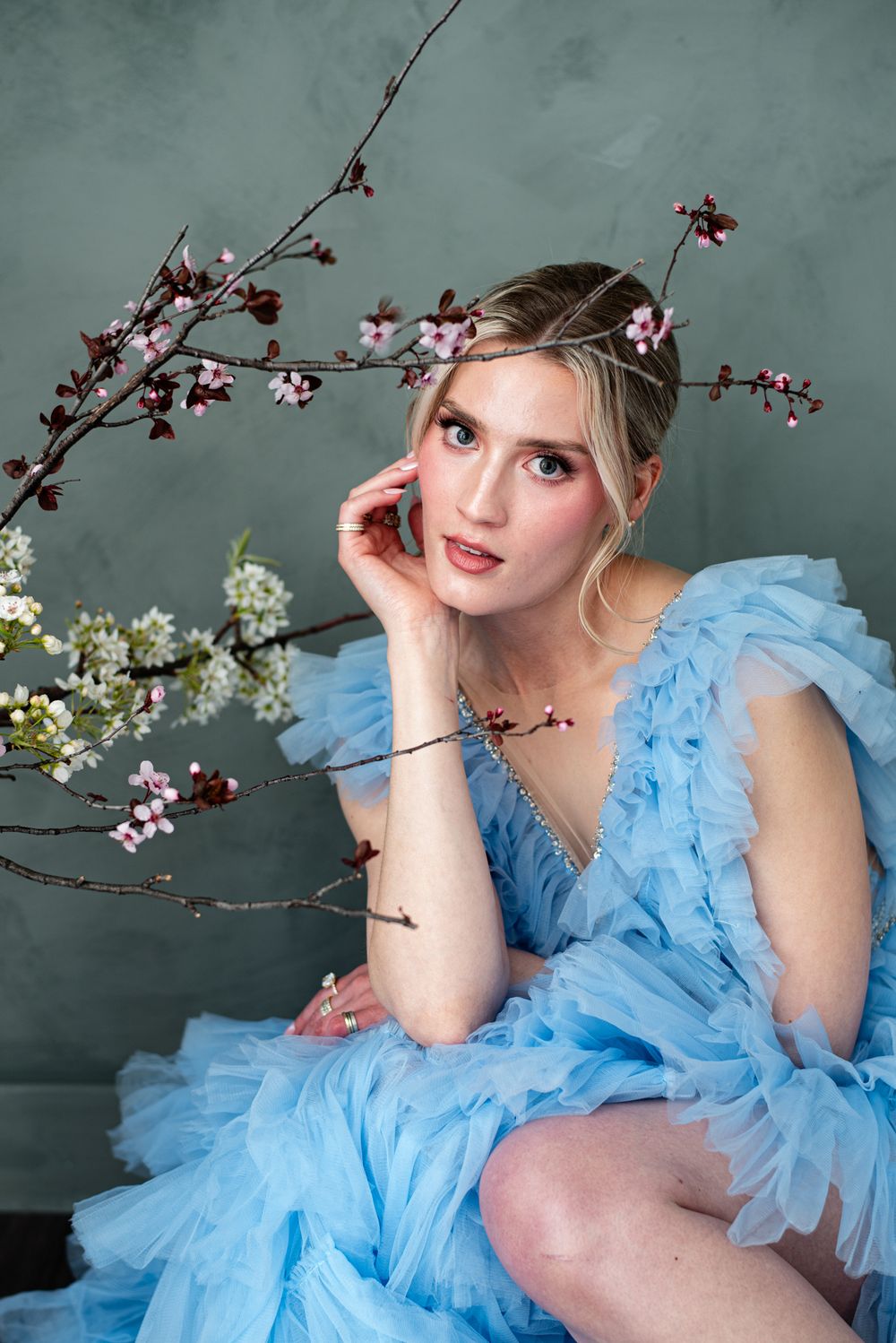 Ethereal portrait in light blue tulle gown posed with delicate cherry blossoms against moody grey backdrop.