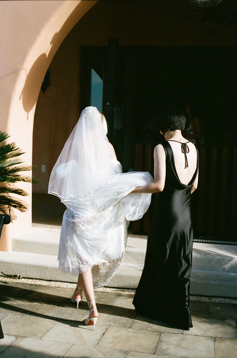 A bride in white dress and bridesmaid in black gown walking together outdoors.