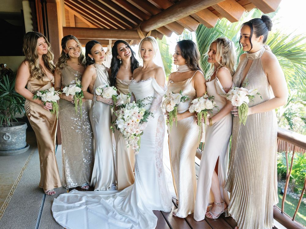 Wedding party poses in champagne-colored dresses with white floral bouquets on a covered patio.