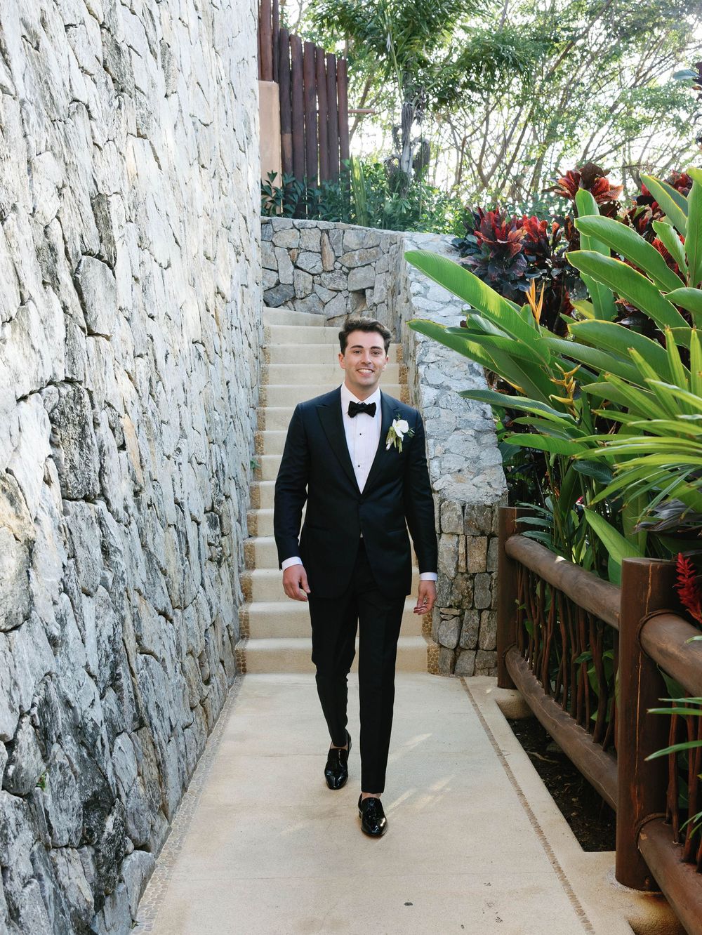 Groom in classic black tuxedo walks along stone-walled pathway with tropical plants.