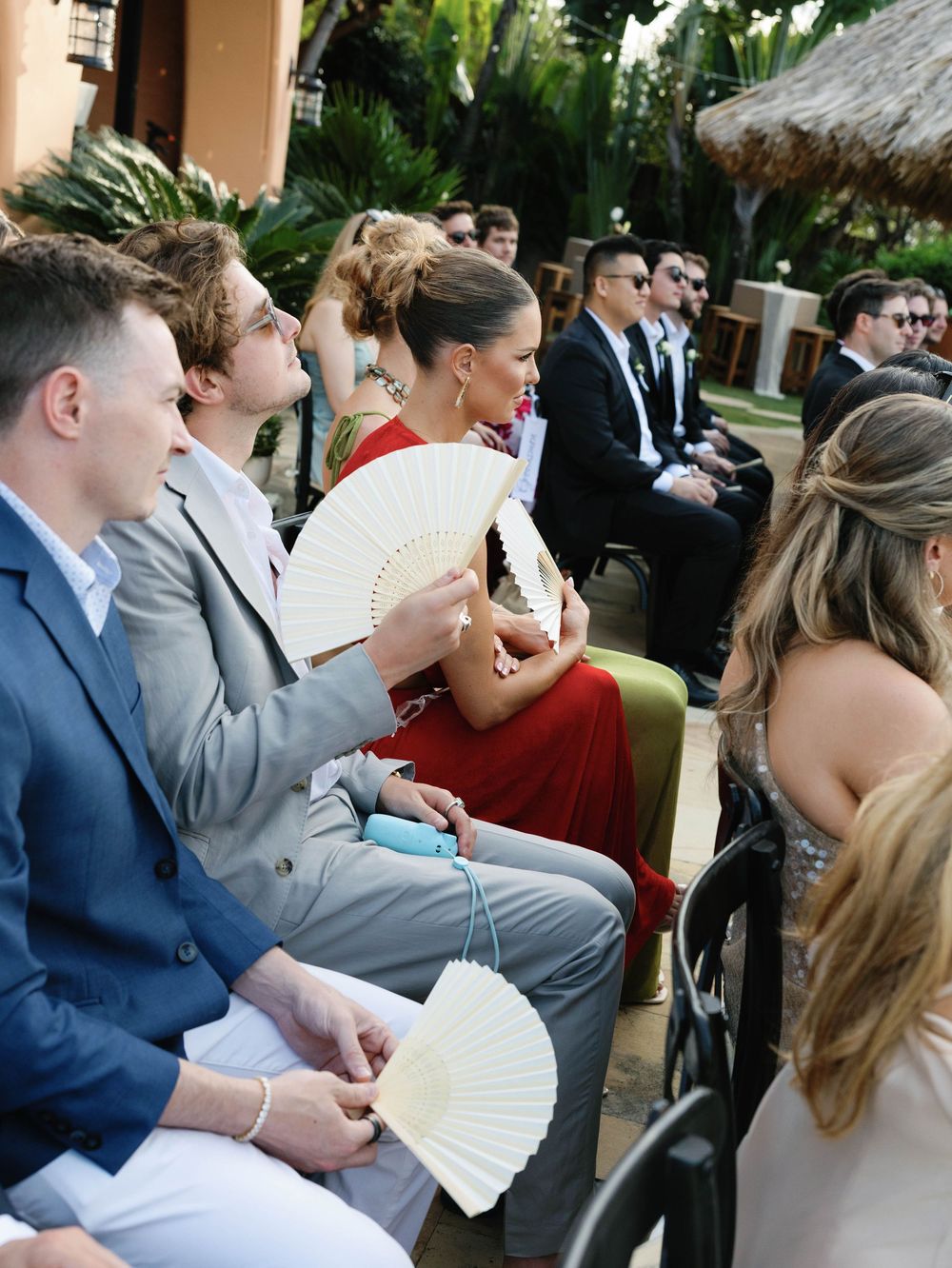 Wedding guests holding white paper fans during an outdoor ceremony in a tropical garden setting.