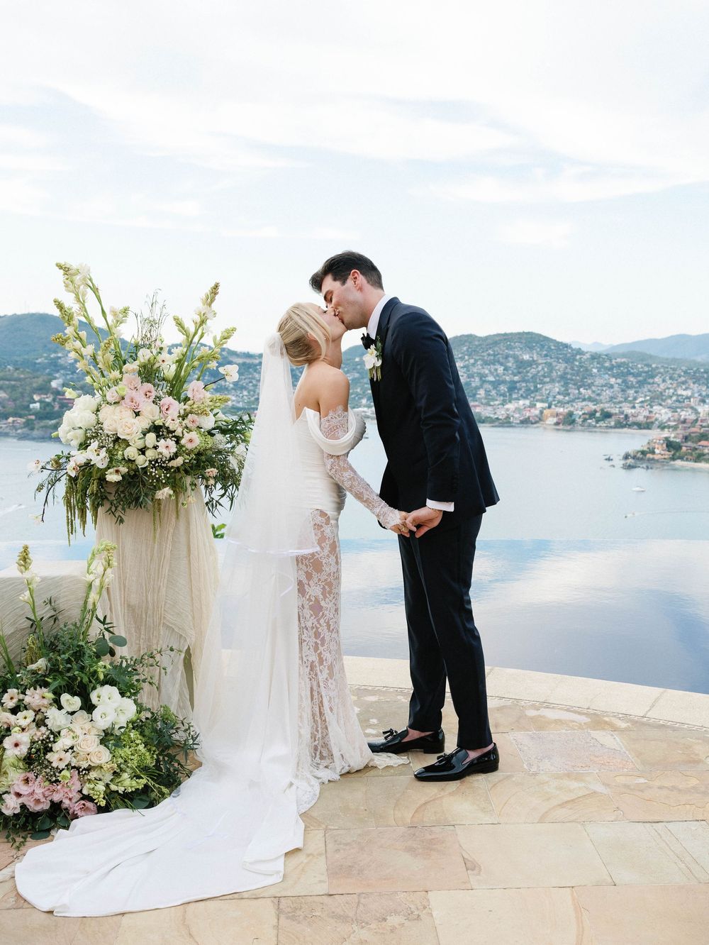 Wedding couple shares intimate moment by floral arch with mountain lake backdrop.