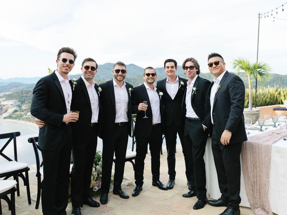 Group of groomsmen in black suits and sunglasses posing on terrace with mountain view.