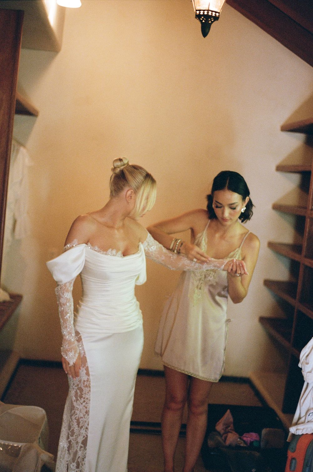 Two women in white preparing final touches of bridal attire in an intimate dressing room moment.