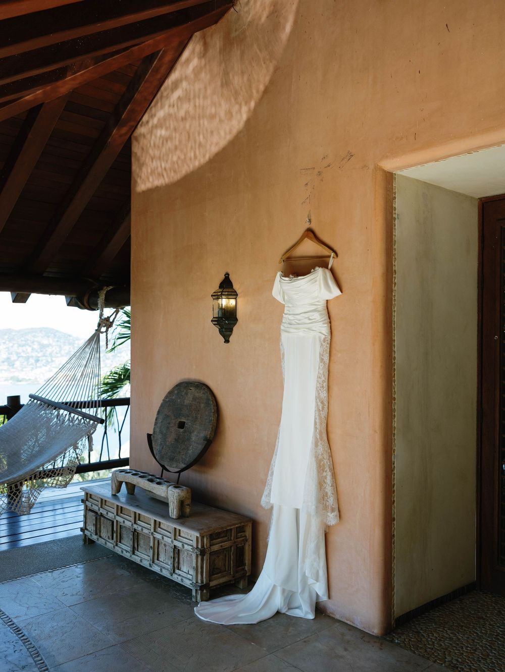 A white wedding dress hangs on display against a beige wall with vintage decor and tropical landscaping visible.