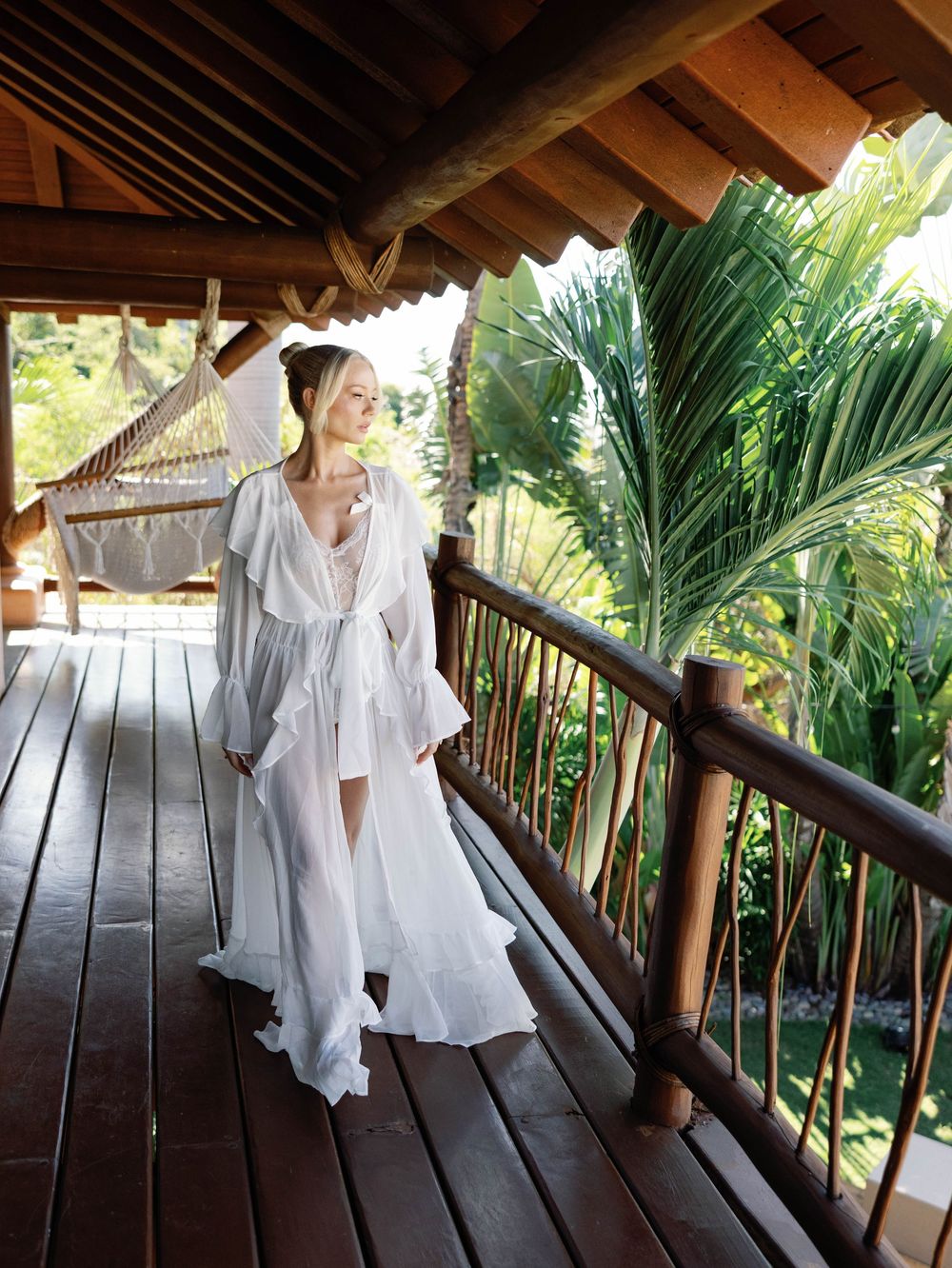 Flowing white robe billows in tropical breeze on wooden balcony surrounded by palm fronds.