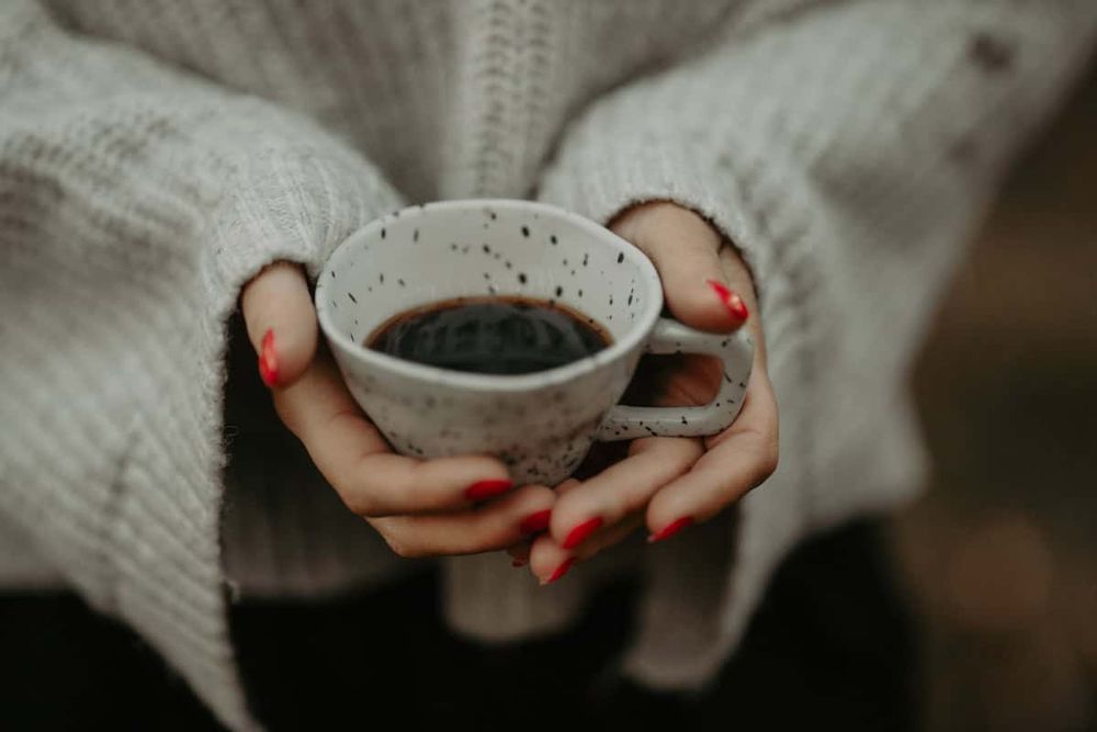 Hands with red nail polish hold a white ceramic mug filled with dark coffee while wearing a cozy knit sweater.