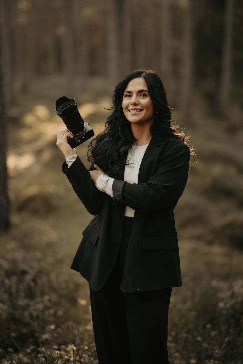 Professional photographer in black blazer holding camera lens in autumn forest with warm lighting.